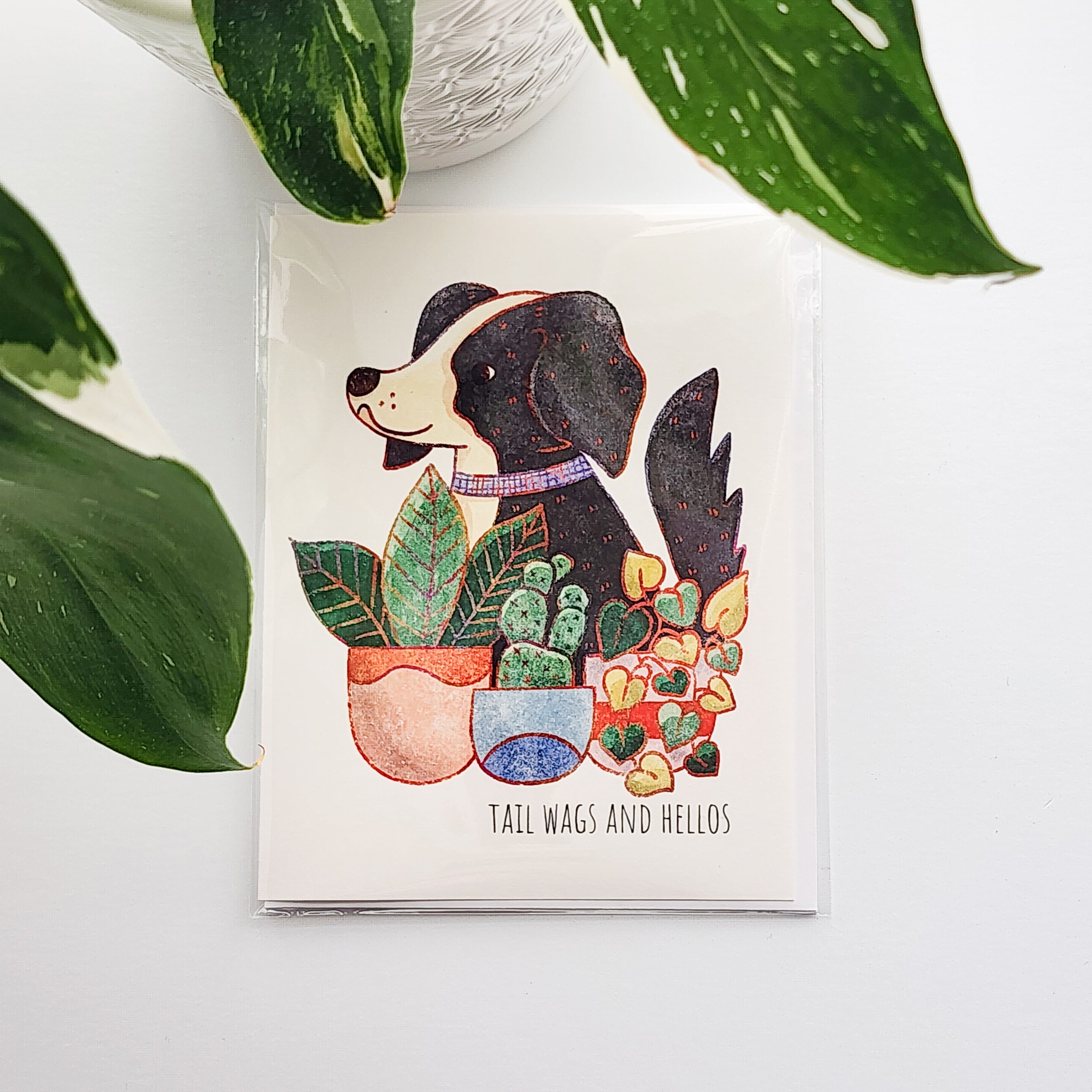 Card with a dog illustration surrounded by plants on a white surface with a potted plant