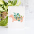 Card with a dog design holding plants, placed on a white surface with a plant in the background.