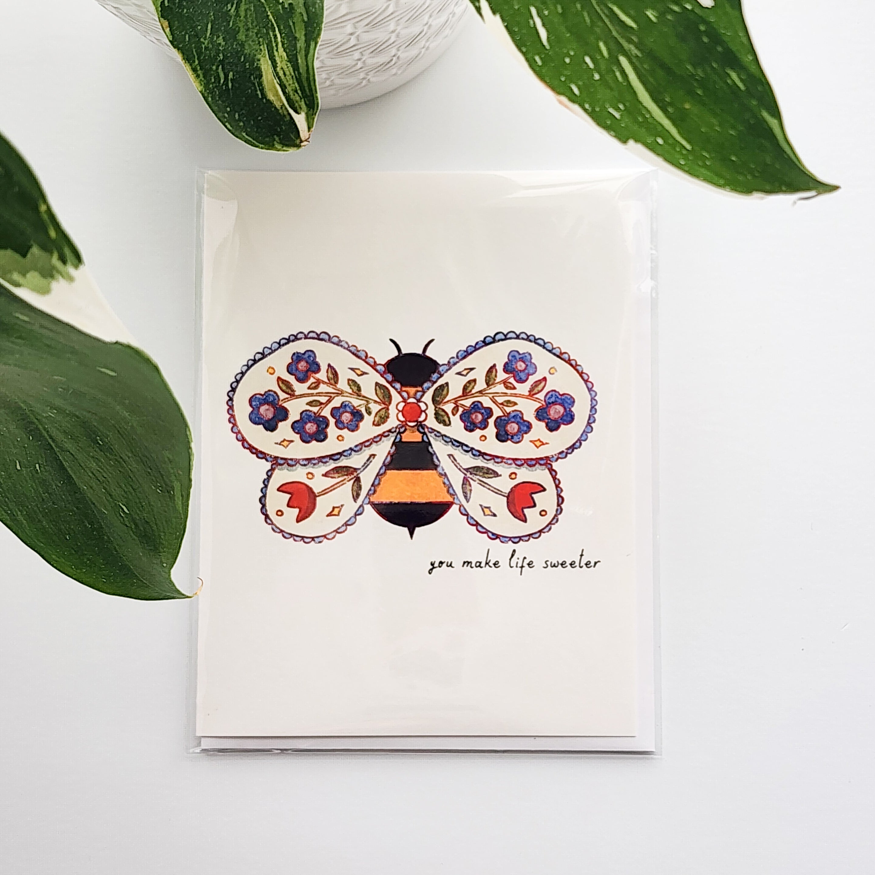 Card with a bee design with flowers on a white surface with a plant in the background