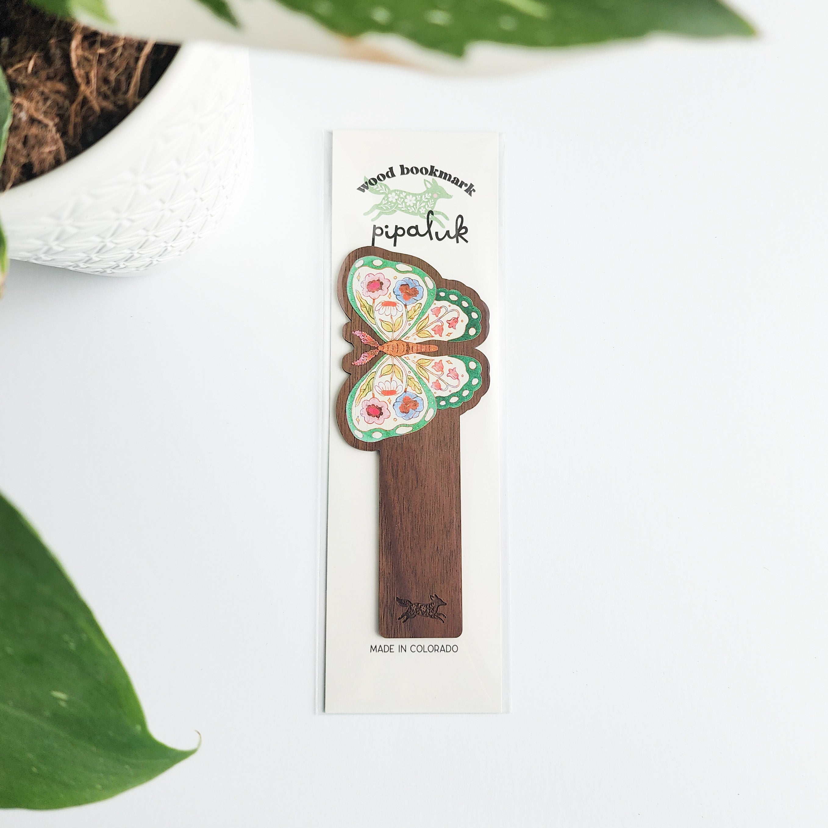 Wooden bookmark with floral design in pipaluk packaging, surrounded by green leaves.