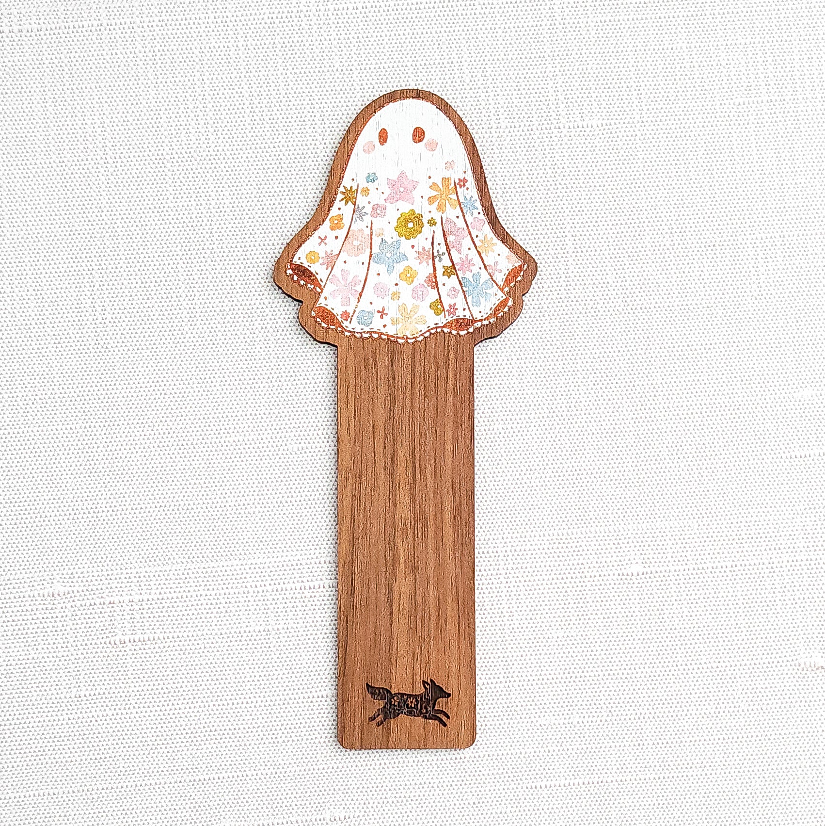 Wooden bookmark featuring a little floral ghost with colorful flowers and stars on its sheet, printed on walnut wood and photographed on a neutral fabric background.