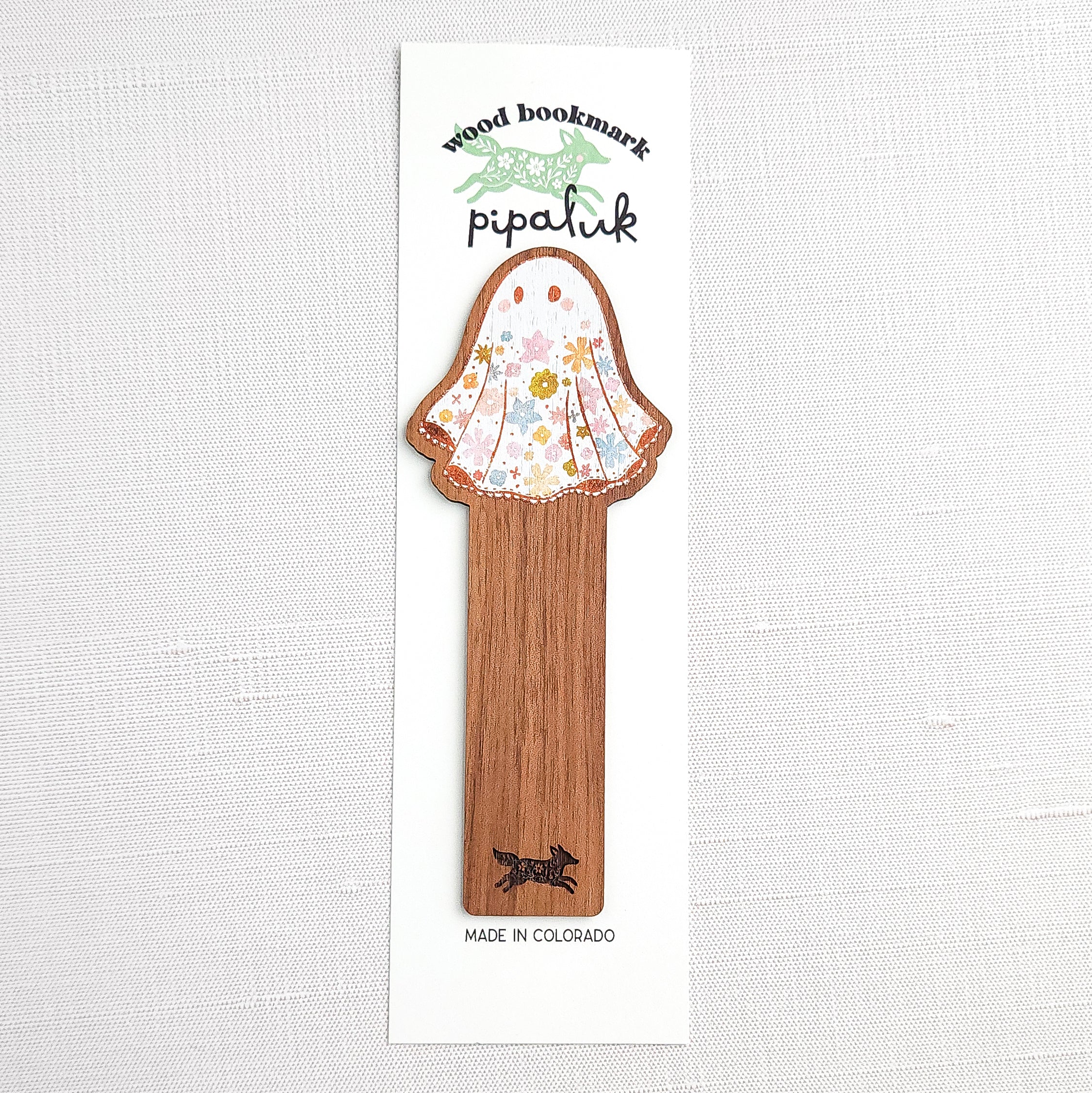 Wooden bookmark featuring a little floral ghost with colorful flowers and stars on its sheet, printed on walnut wood and photographed in Pipaluk branded packaging on a neutral fabric background
