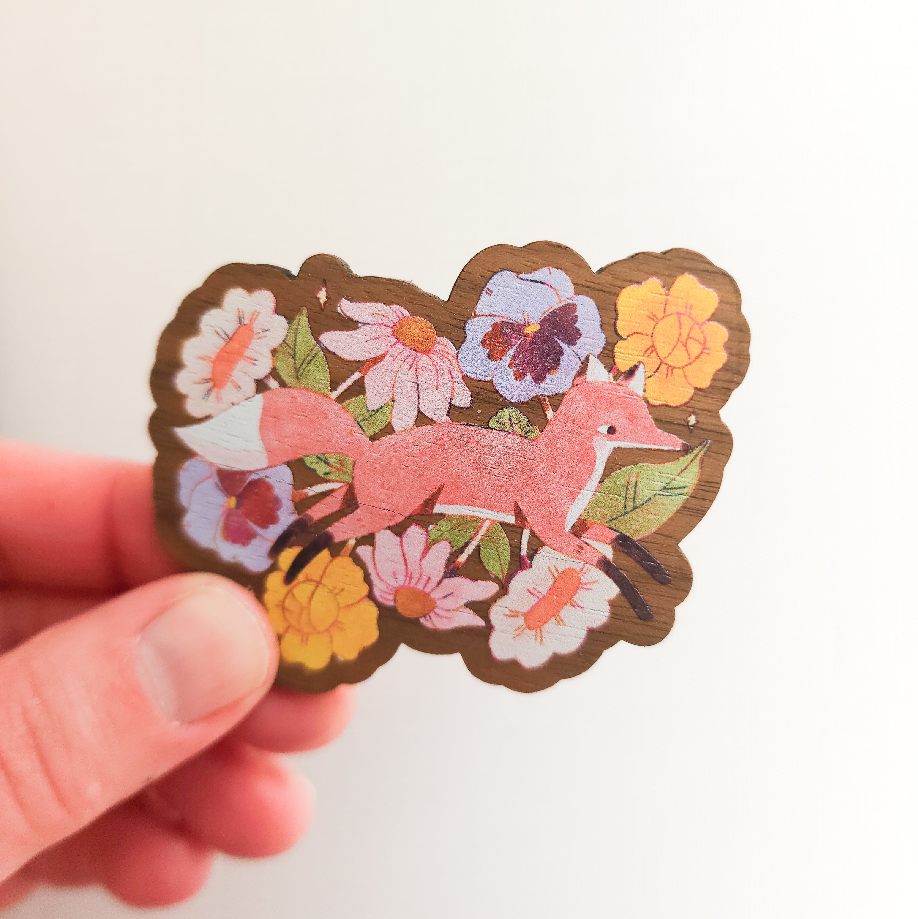 hand holding a wooden magnet with an illustration of a fox jumping through flowers on a linen background