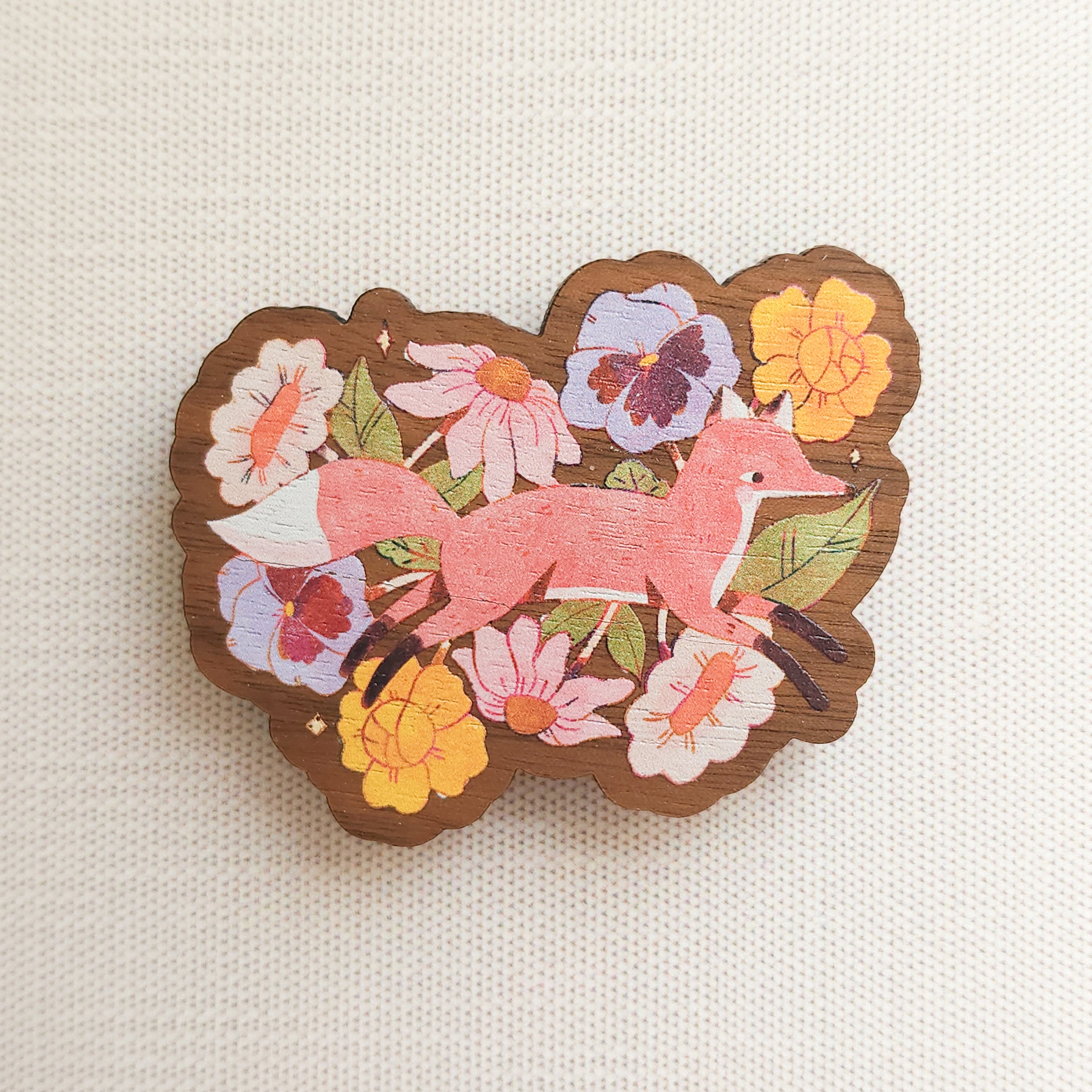 wooden magnet with an illustration of a fox jumping through flowers on a linen background