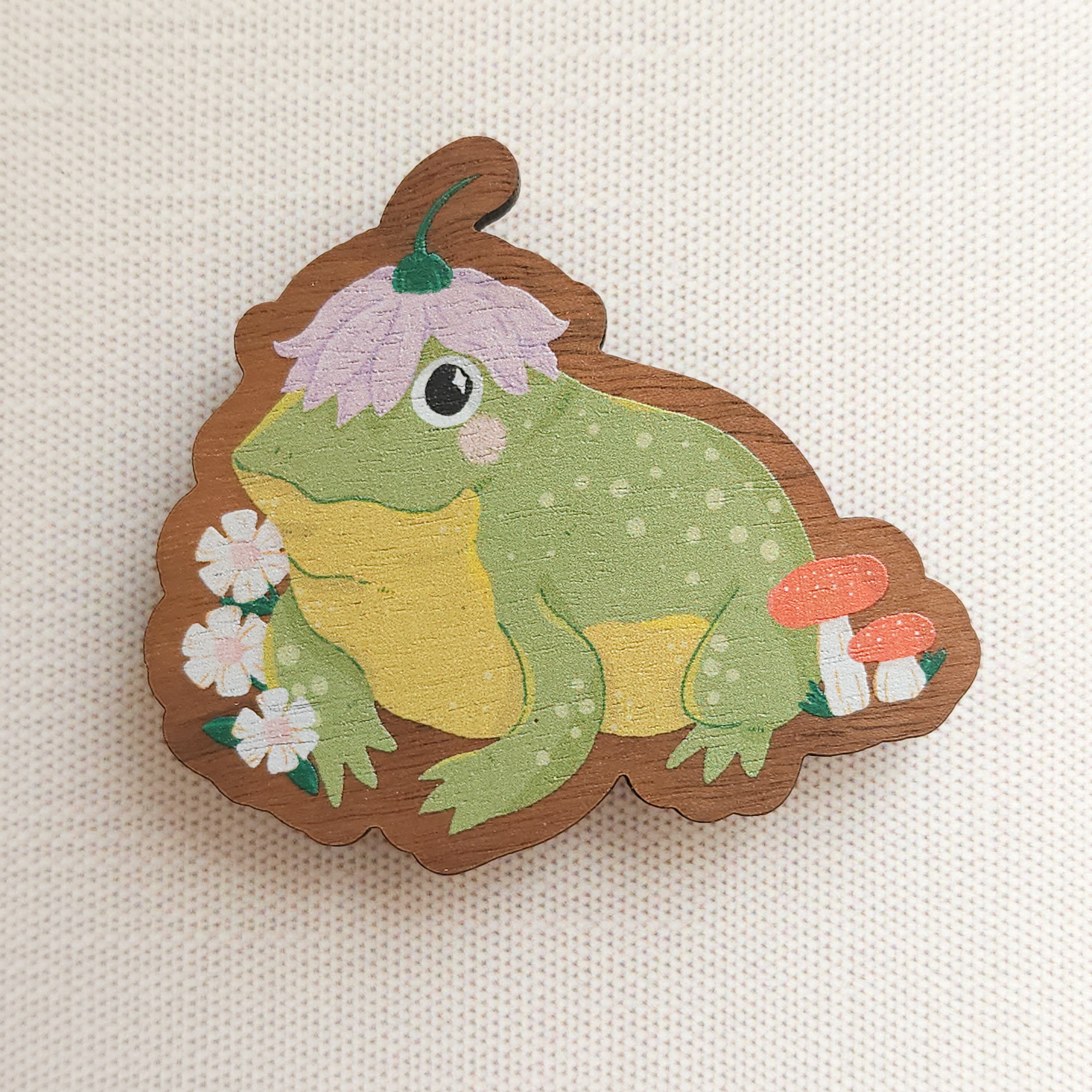 wooden magnet with an illustration of a happy toad wearing a flower a hat on a linen background