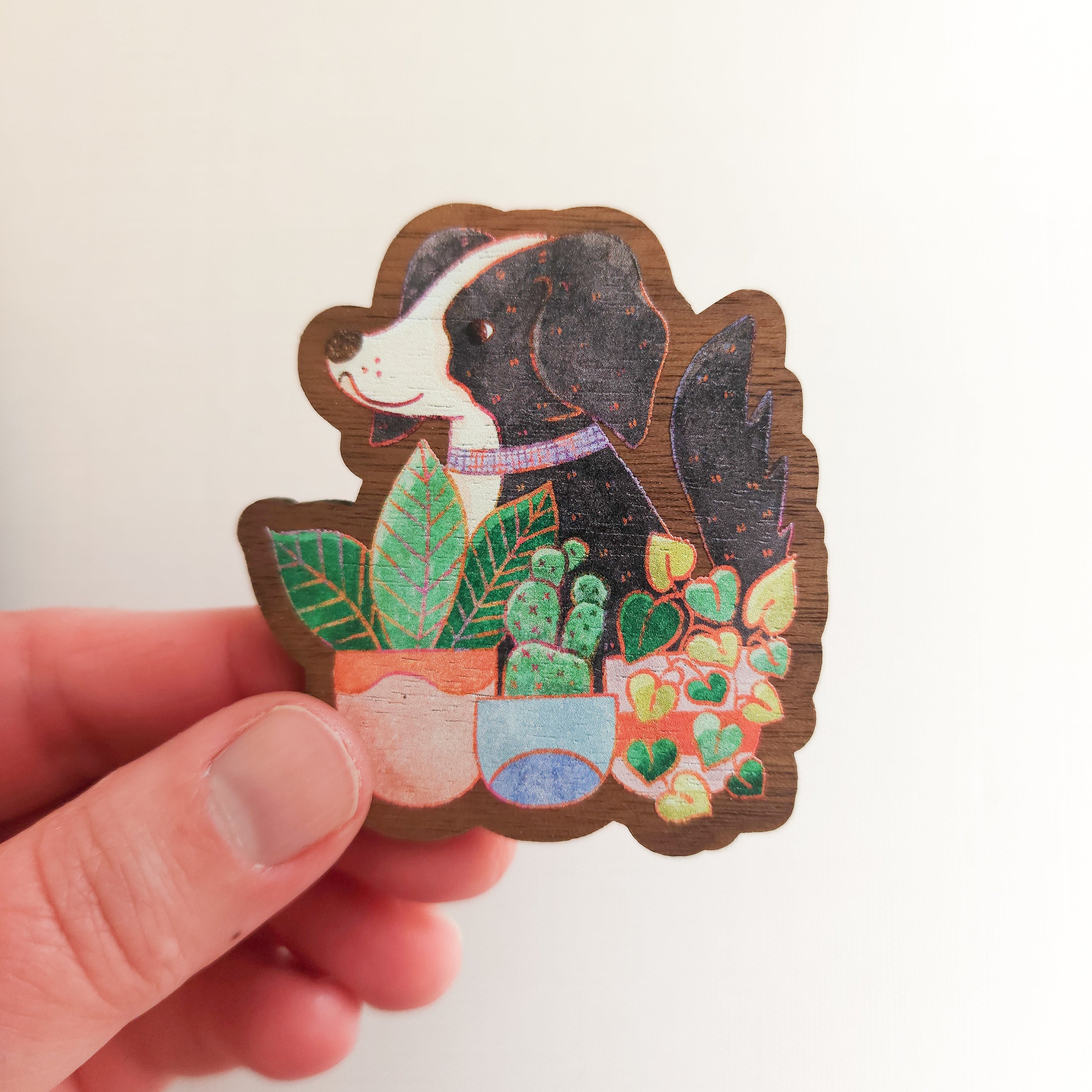 hand holding a wooden magnet with an illustration of a black and white dog sitting behind potted houseplants on a linen background