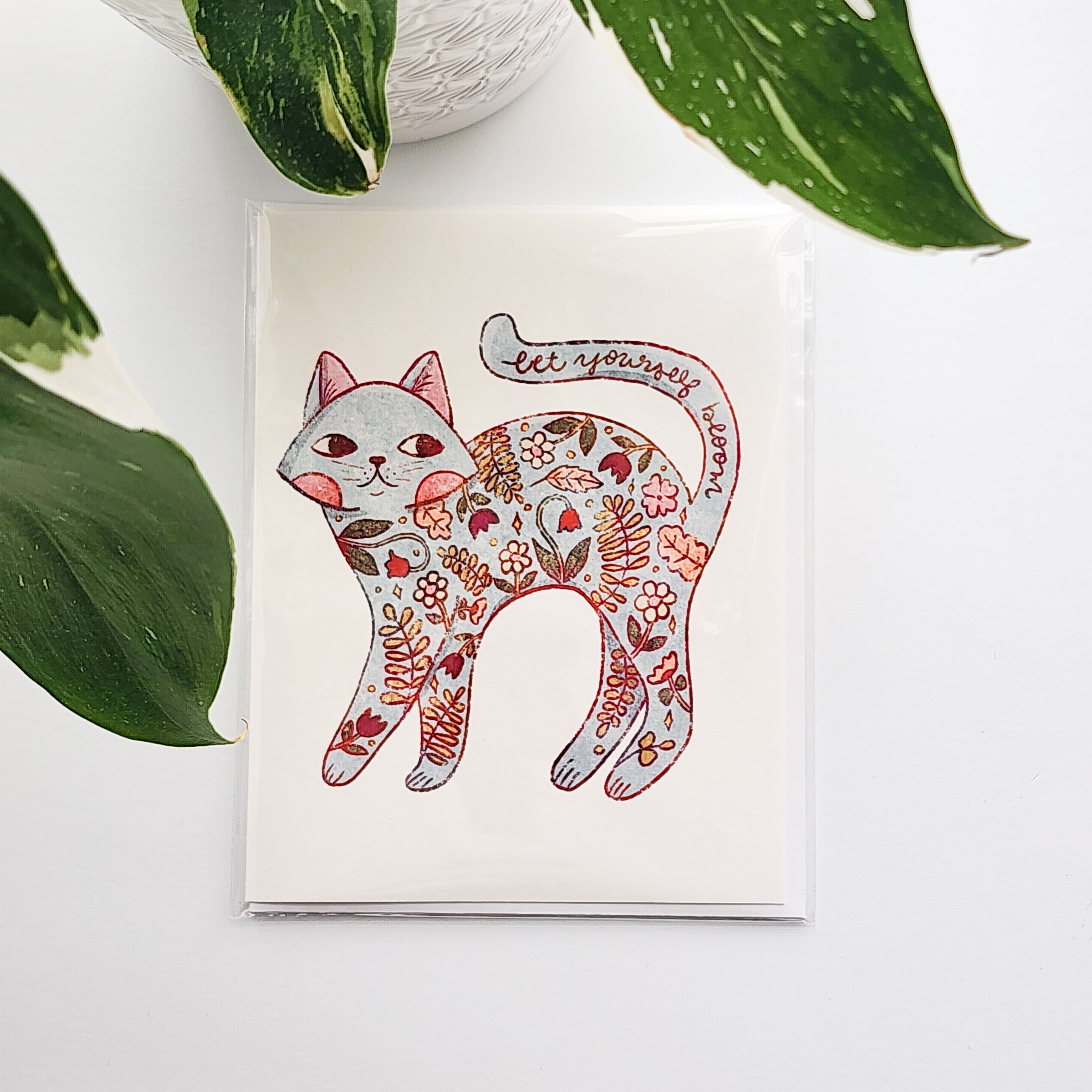 card with an illustrated floral cat and the words let yourself bloom on a white background in front of a plant
