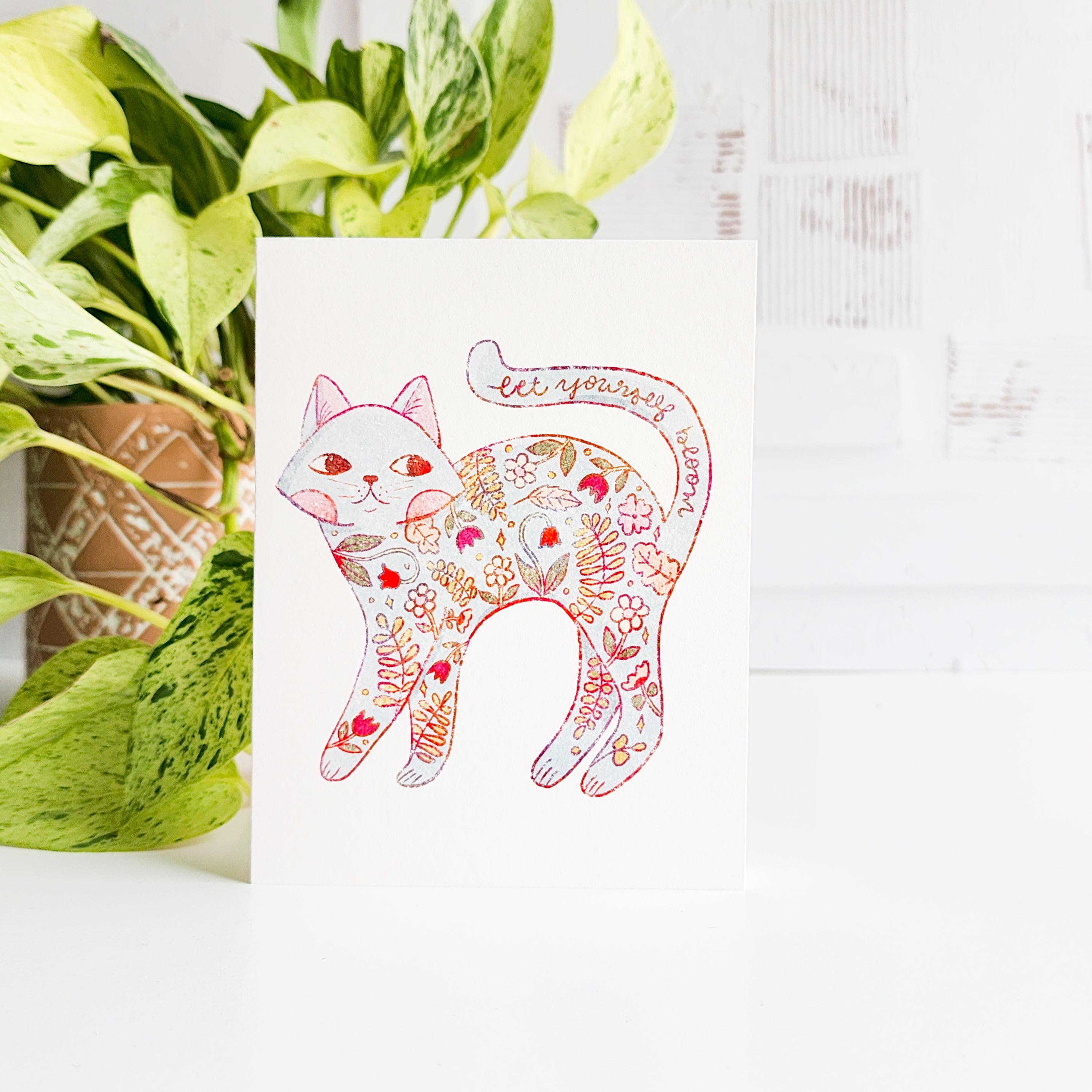 Decorative card with a floral cat design next to a potted plant on a white surface.