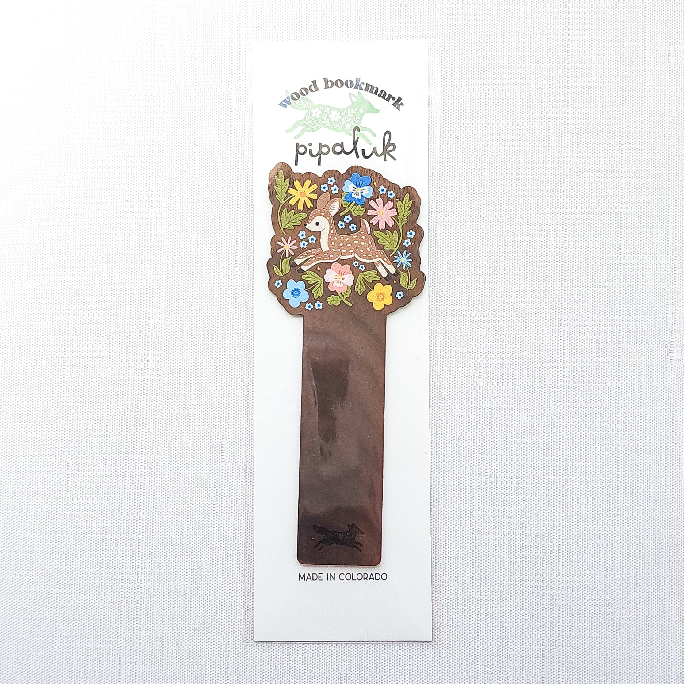 Wooden bookmark featuring a sweet spotted fawn dancing among colorful wildflowers, printed on walnut wood and photographed in Pipaluk packaging on a neutral fabric background