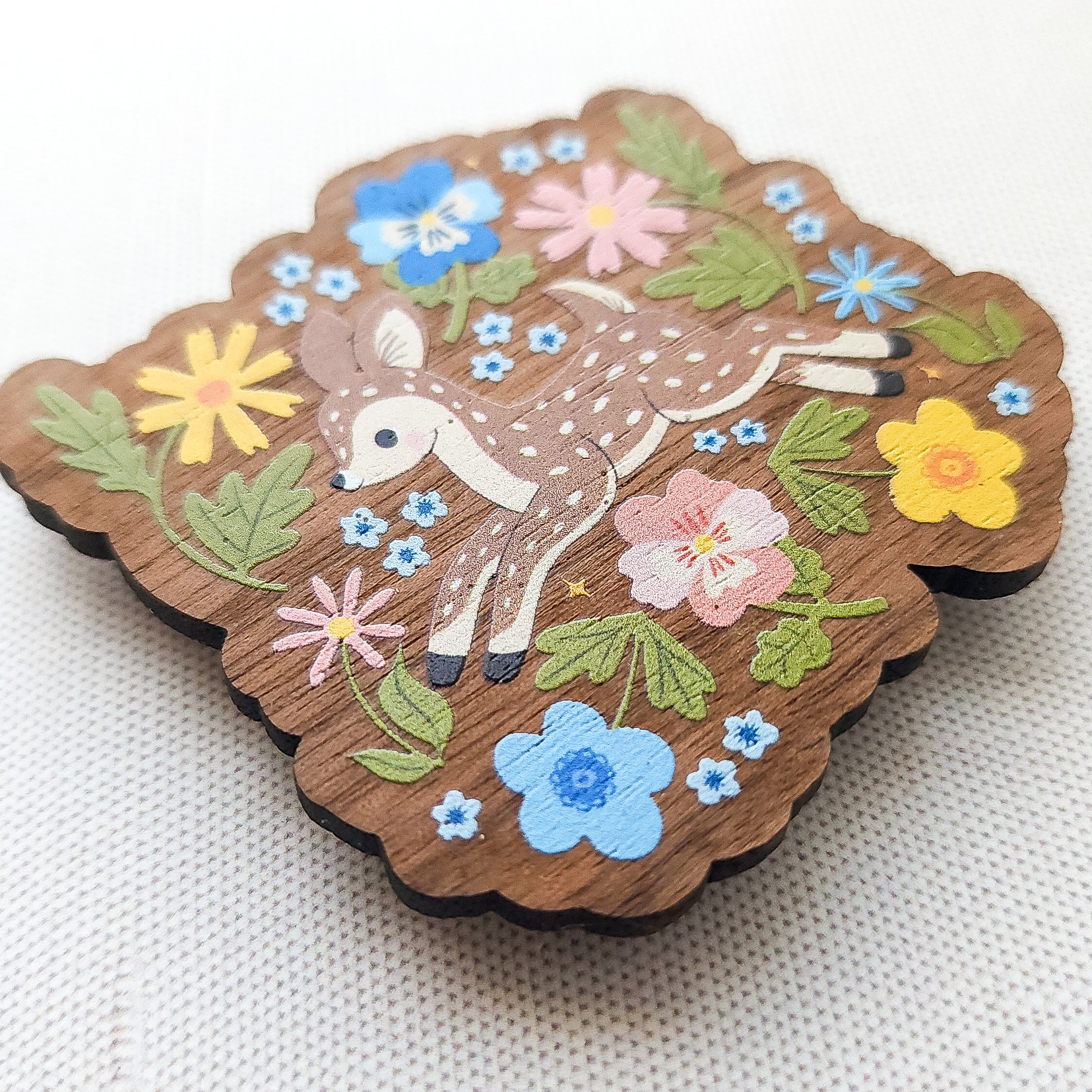 Wooden magnet featuring a sweet spotted fawn dancing among colorful wildflowers, printed on walnut wood and photographed on a neutral fabric background.