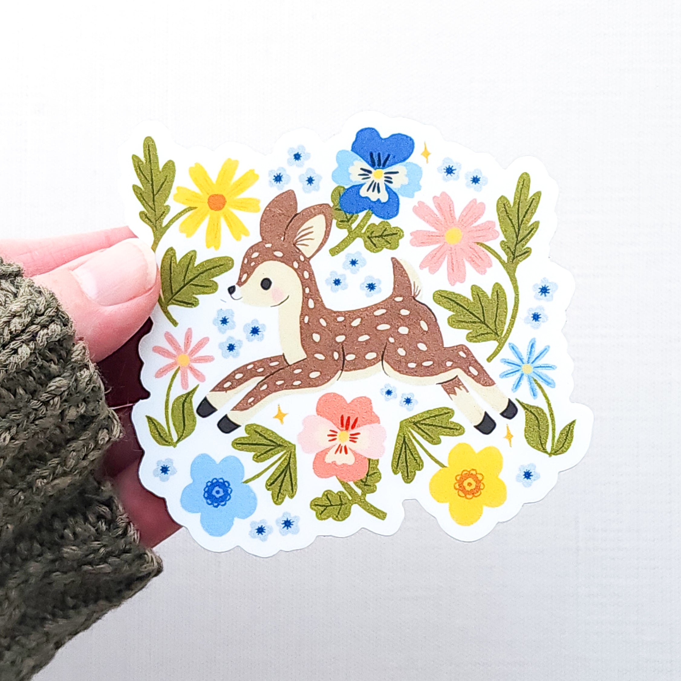 Vinyl sticker featuring a sweet spotted fawn dancing among colorful wildflowers, with pink, yellow, blue, and green floral details held in hand on a neutral background.