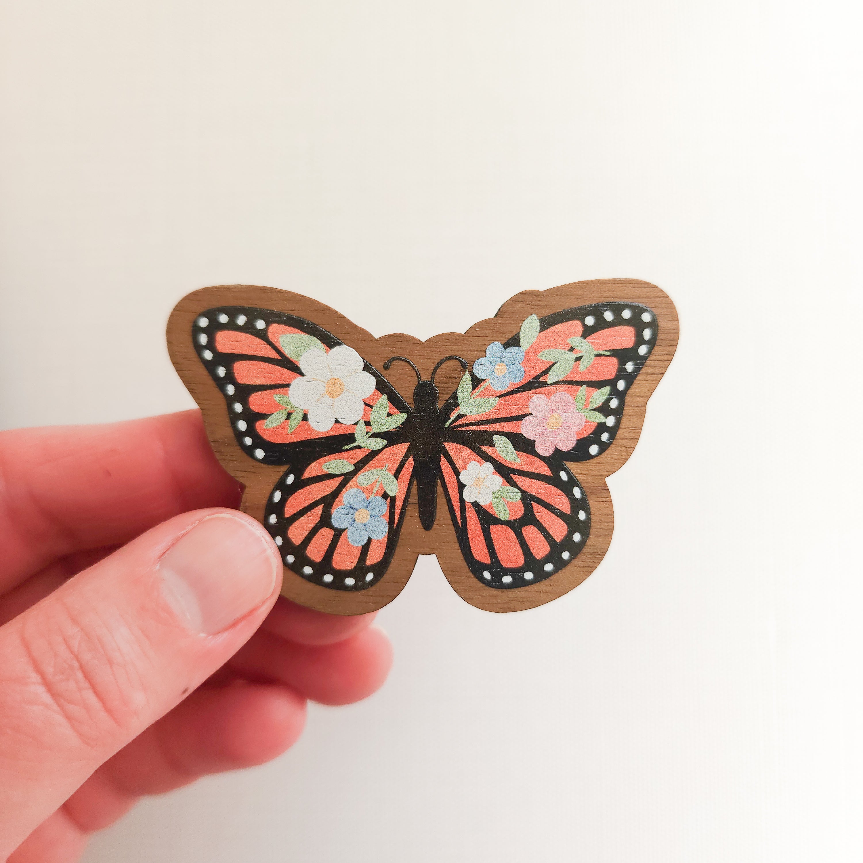 hand holding a wooden magnet with an illustration of a monarch butterfly with flowers in its wings