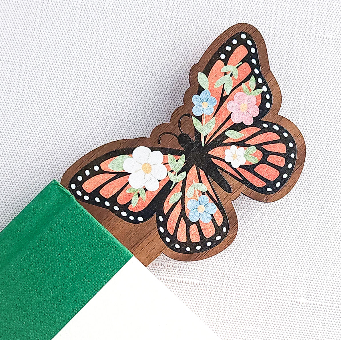 Wooden bookmark featuring a monarch butterfly with orange wings, black detailing, and tiny colorful blooms, printed on walnut wood and photographed inside of a book on a neutral fabric background.