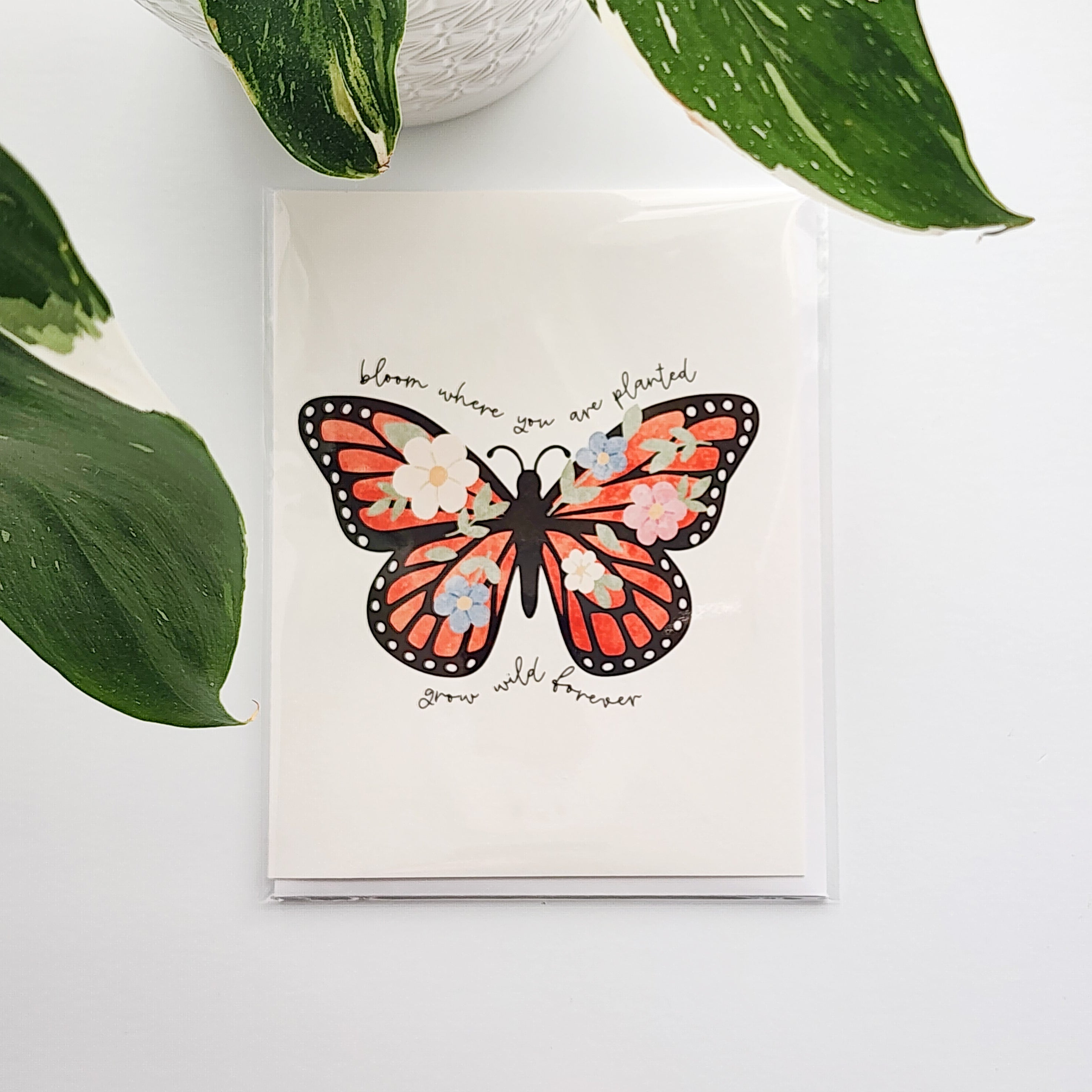 greeting card with an illustrated butterfly and the words bloom where you are planted, grow wild forever. The card is on a white background next to a potted plant
