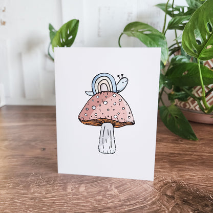 rainbow snail sitting on a mushroom greeting card sitting in front of a plant