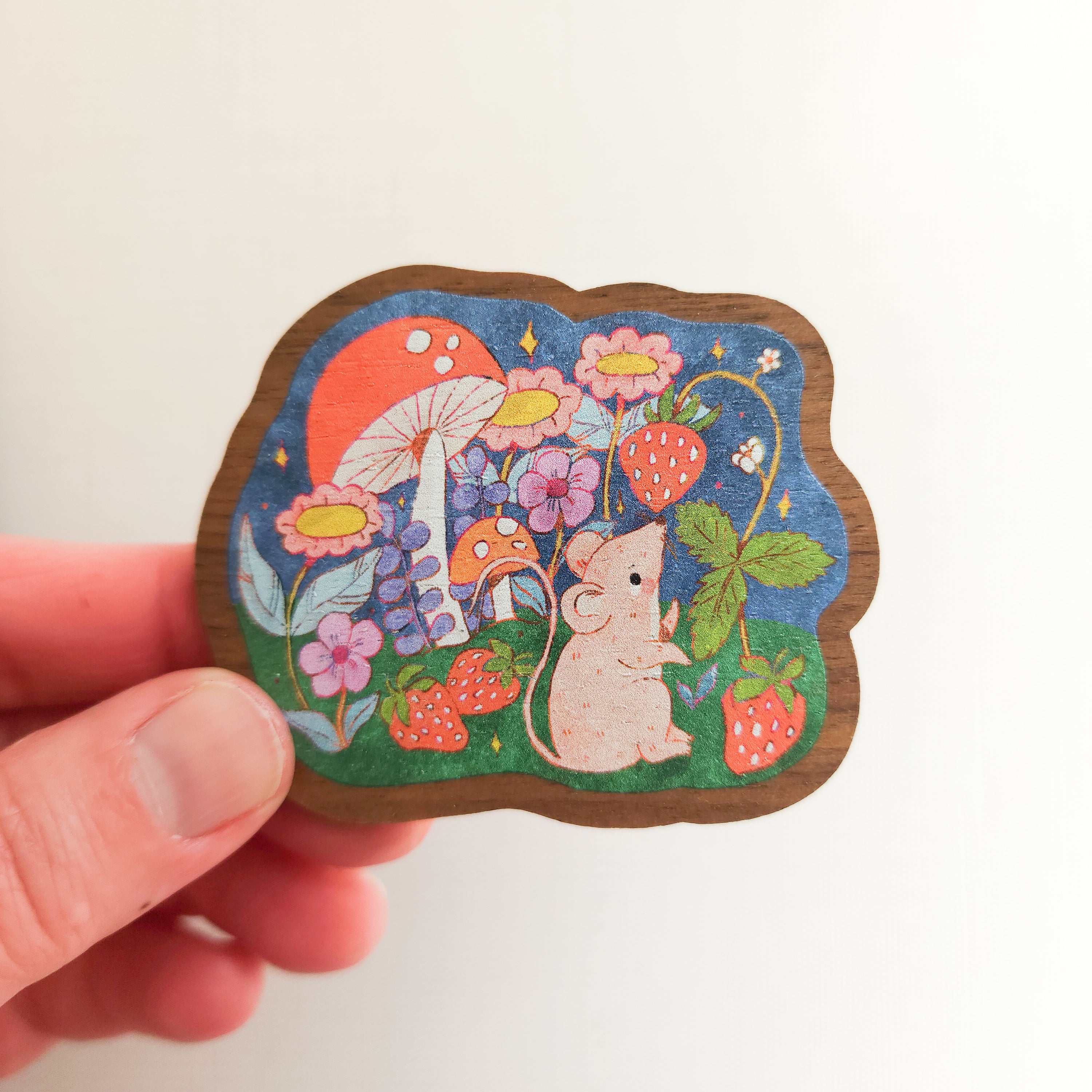 wooden magnet with an illustration of a mouse picking strawberries on a linen background held in a hand