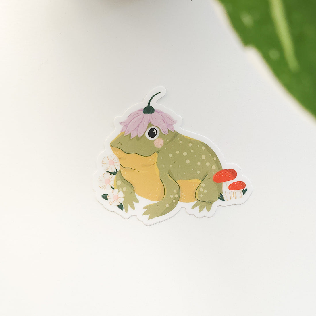 Sticker of a frog with a flower on its head and mushrooms nearby, on a white background.