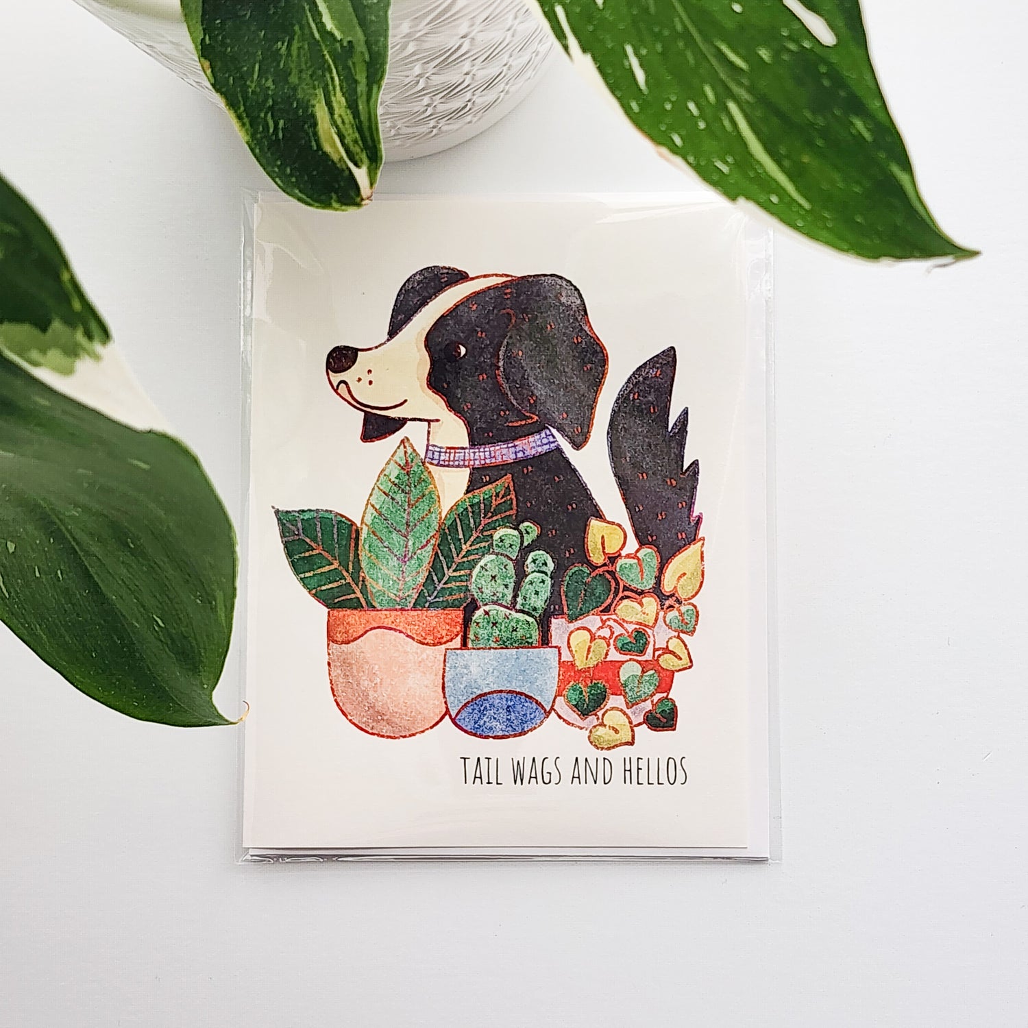 Card with a dog illustration surrounded by plants on a white surface with a potted plant