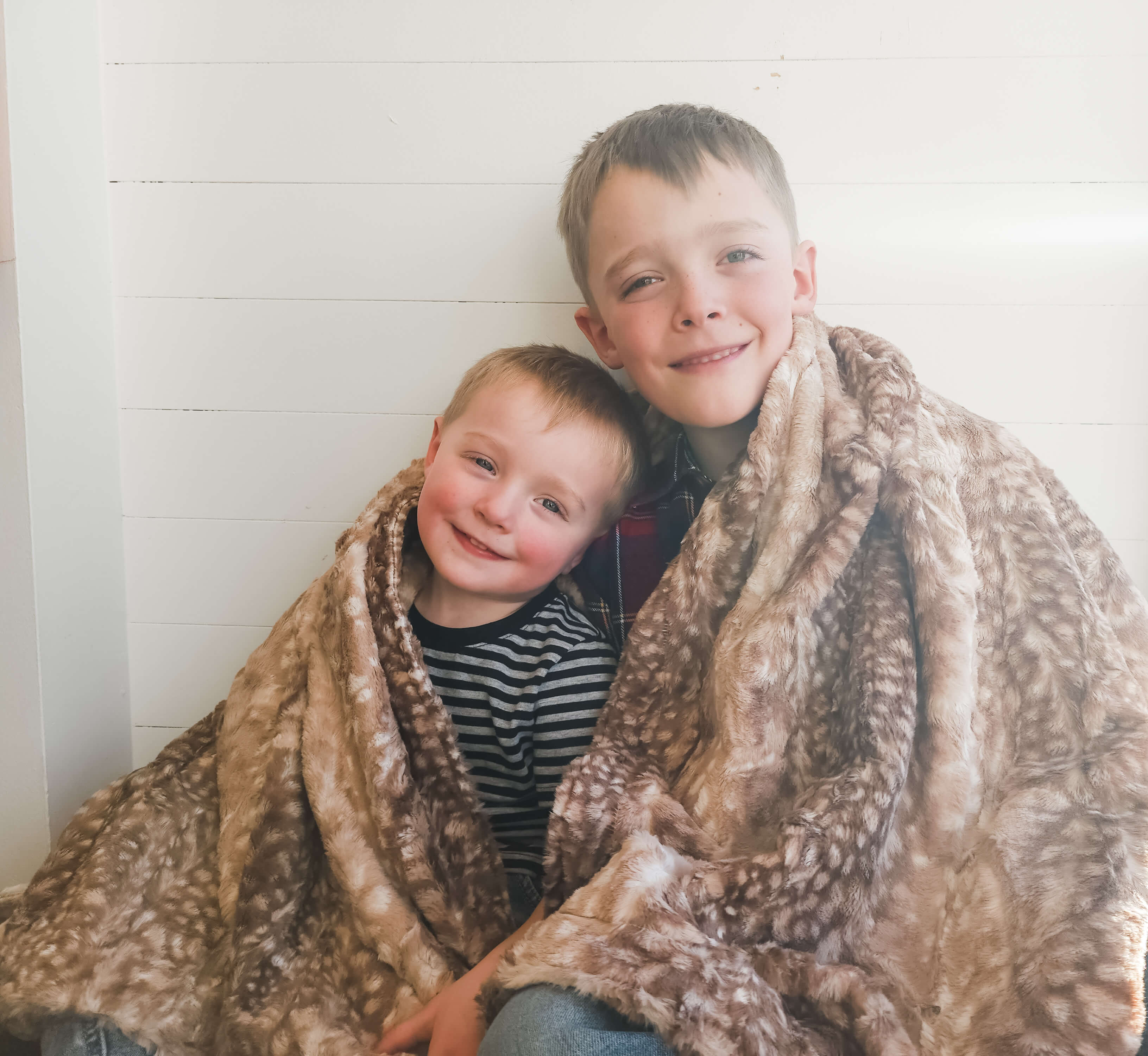 two young boys smiling sitting close together wrapped in a fawn print blanket
