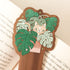 wooden bookmark with an illustration of a cat in a monstera plant sitting on an open book