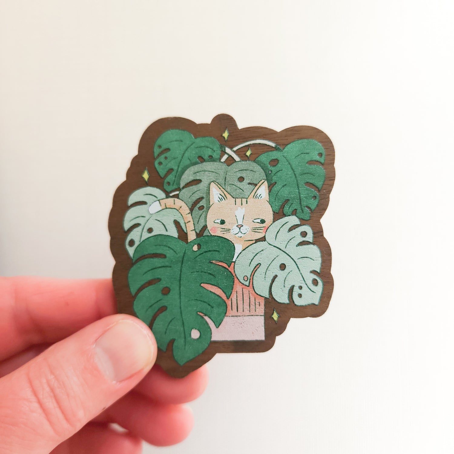 hand holding a wooden magnet with an image of a cat in a monstera plant on a linen background