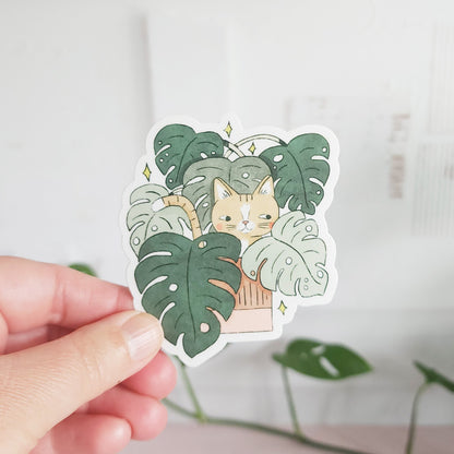 A hand holding a vinyl sticker with an illustration of a cat peeking out from behind monstera leaves.