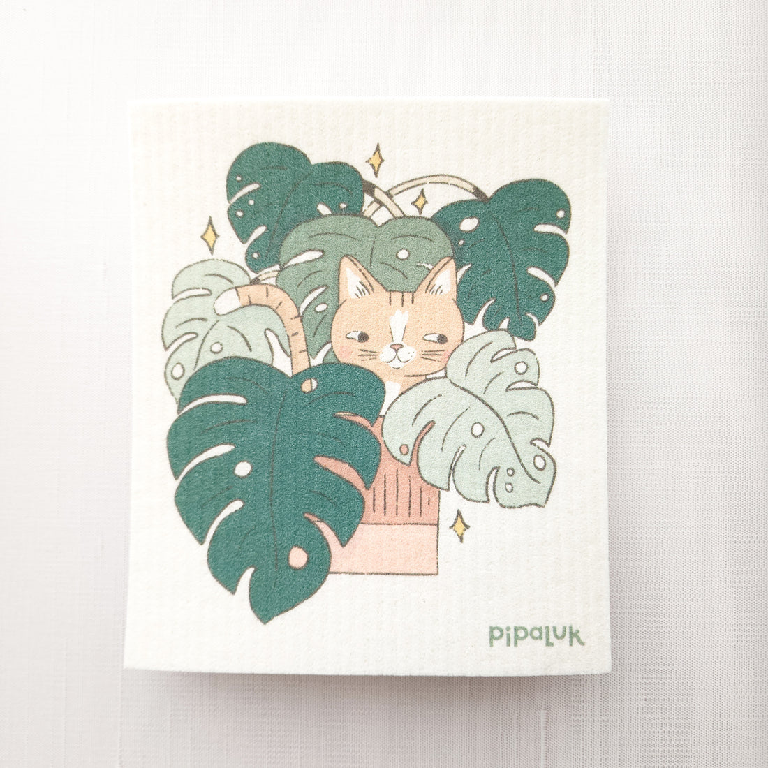 swedish dishcloth with an illustration of a cat in a monstera plant and  pipaluk logo in the bottom right corner
