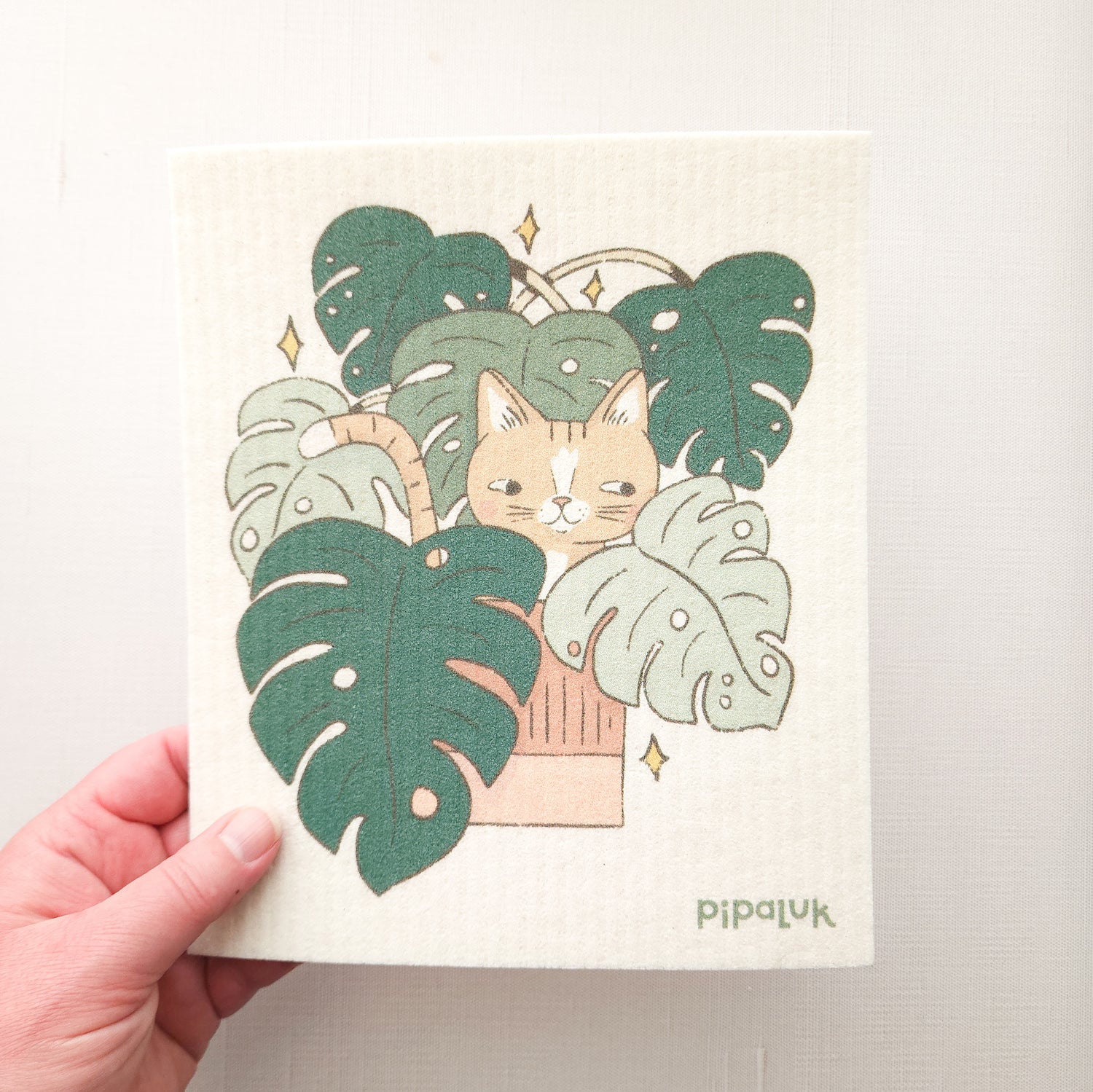 swedish dishcloth with the illustration of a cat in a monstera plant, held in a hand