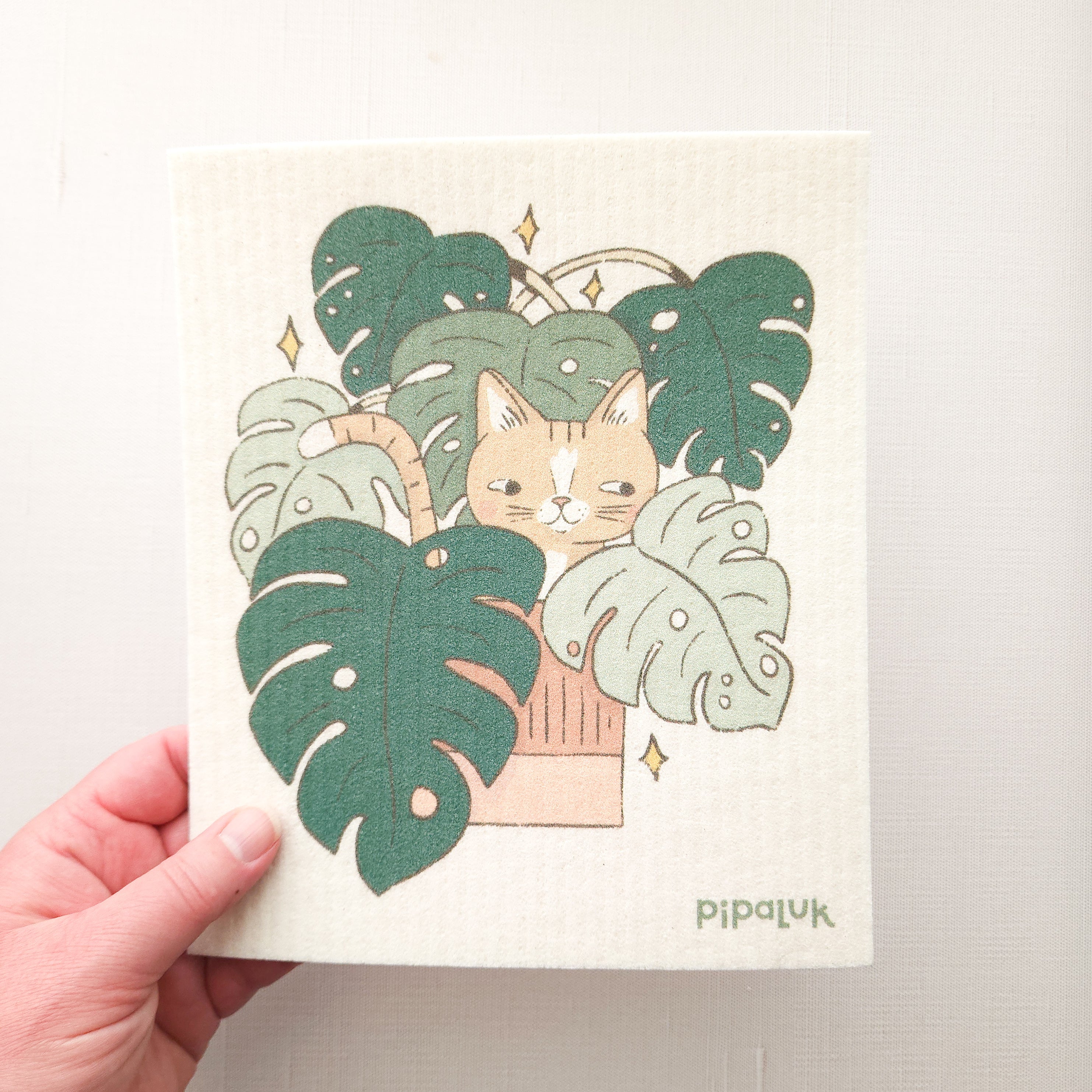 swedish dishcloth with the illustration of a cat in a monstera plant, held in a hand