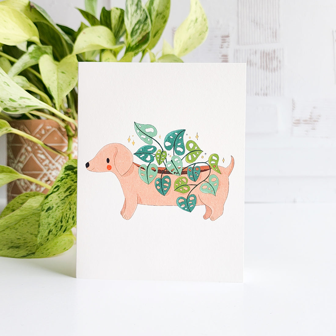 Card with a dog design holding plants, placed on a white surface with a plant in the background.