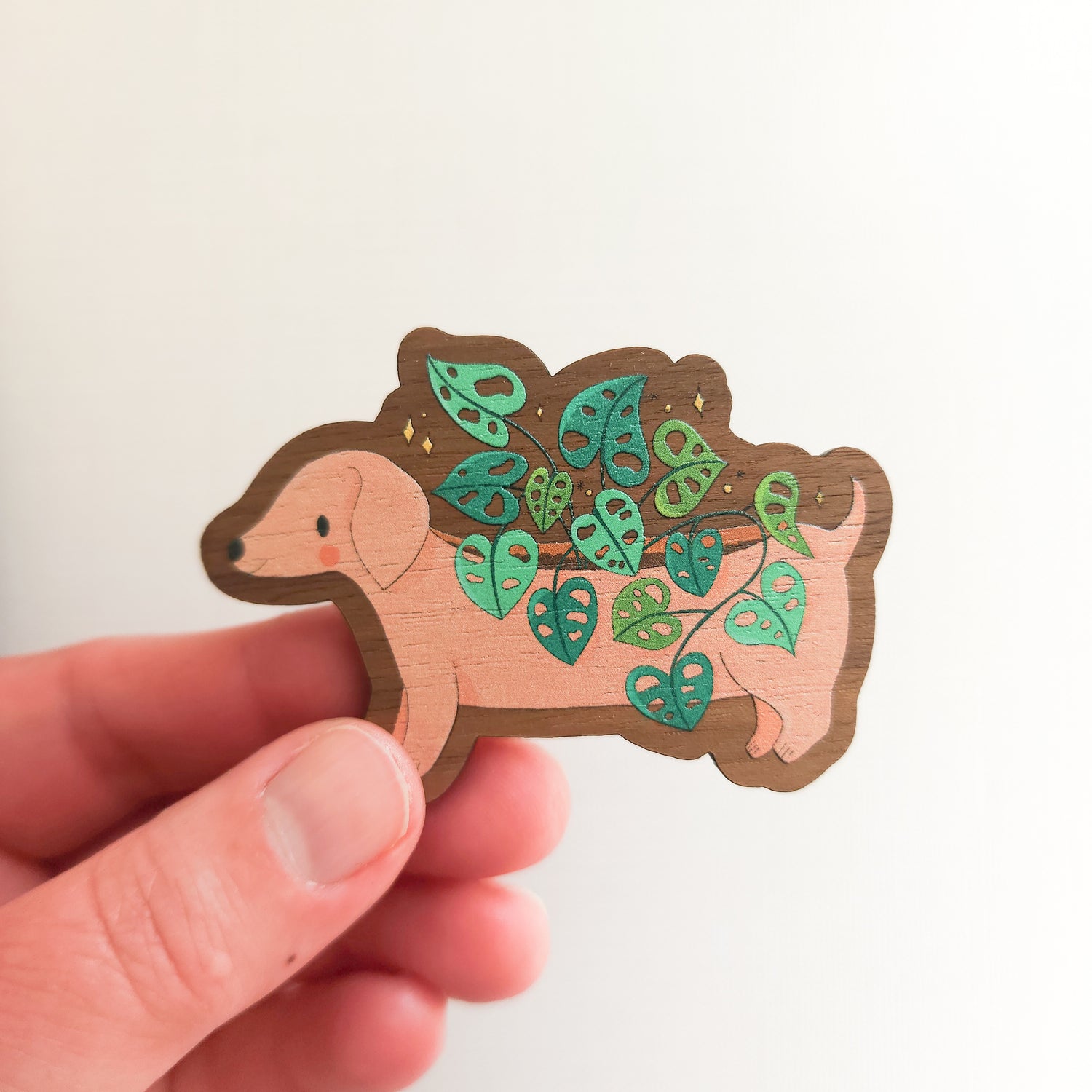 hand holding a wooden magnet with an illustration of a dachshund planter with a cheese plant on a linen background