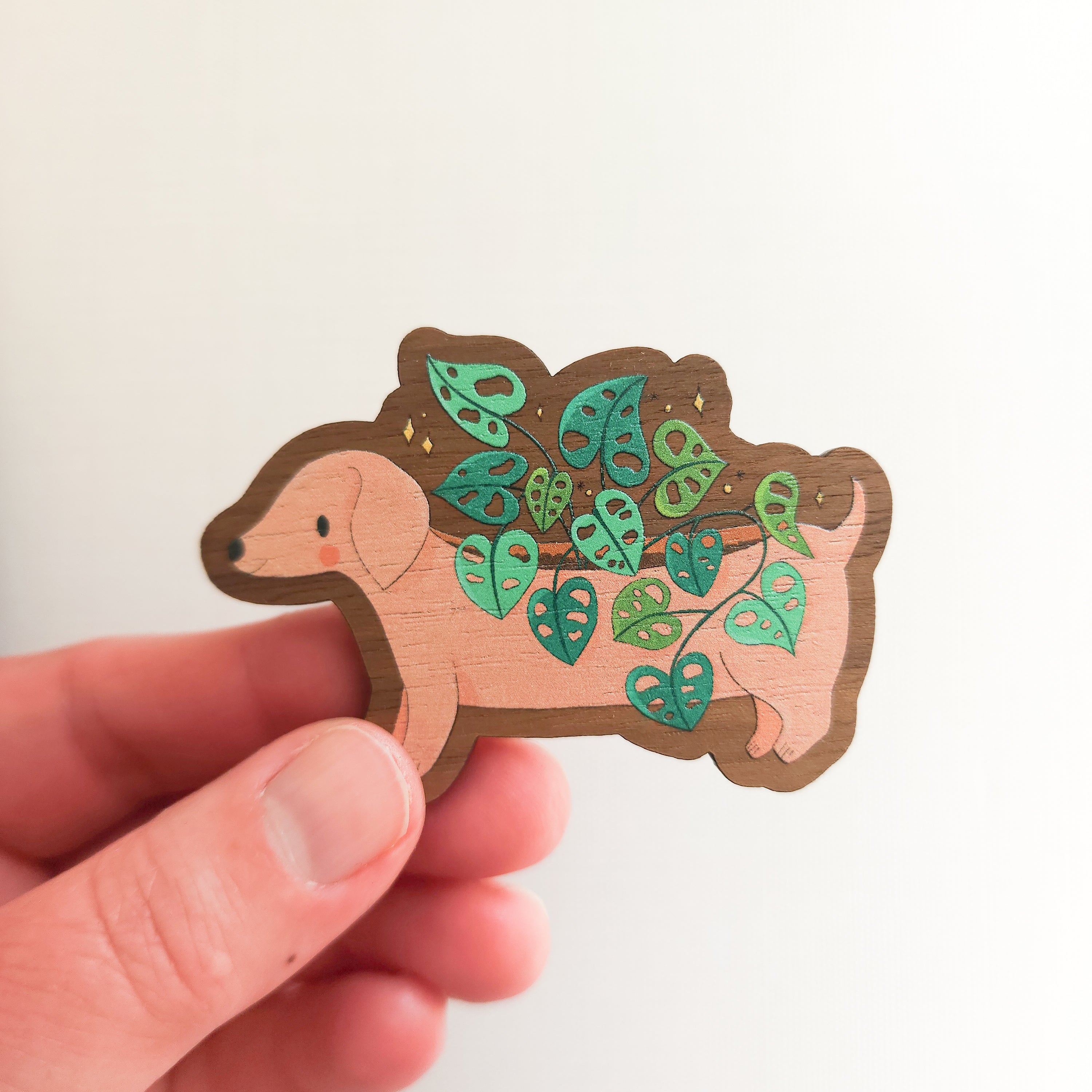 hand holding a wooden magnet with an illustration of a dachshund planter with a cheese plant on a linen background