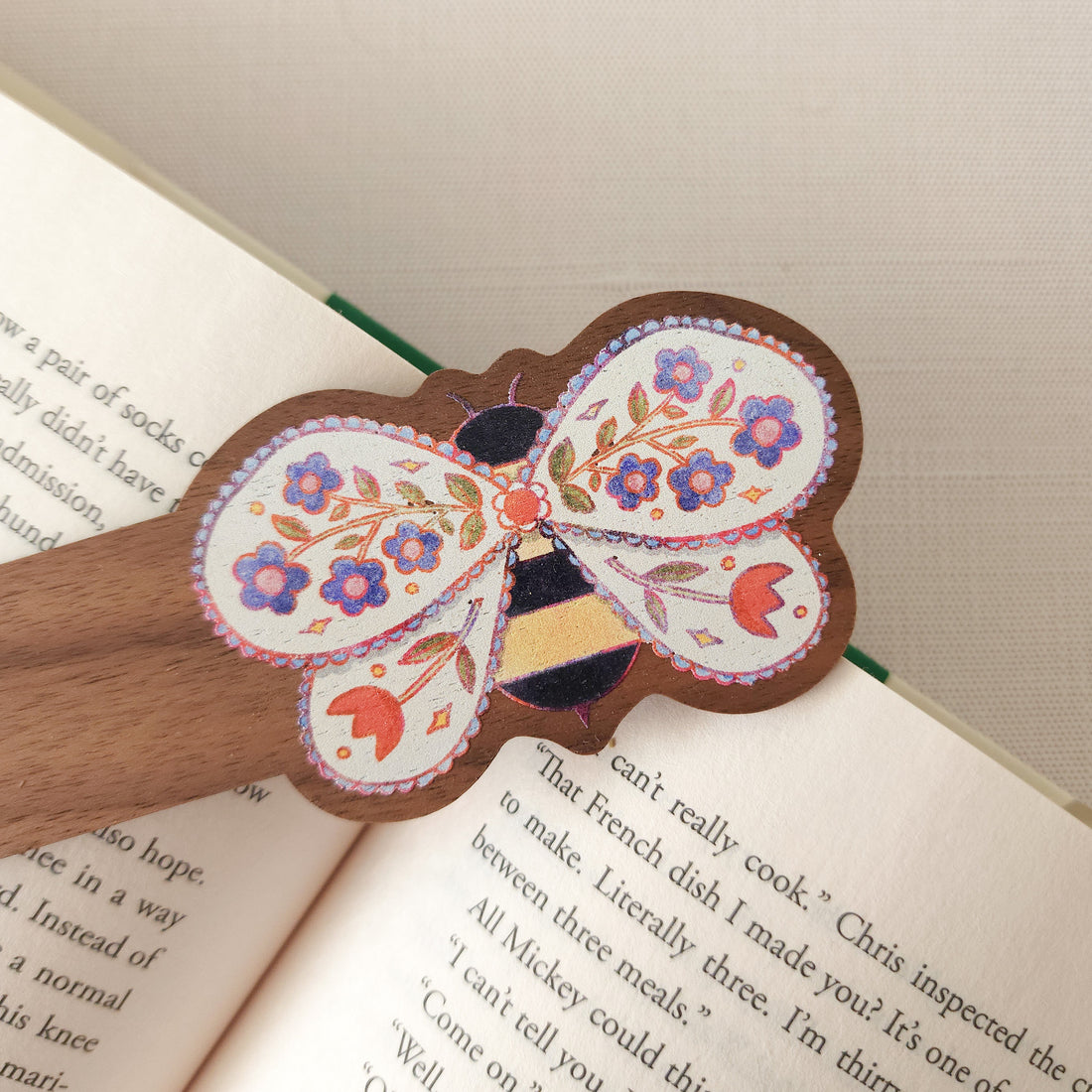wooden bookmark with an illustration of a bee with flowers in its wings sitting on an open book