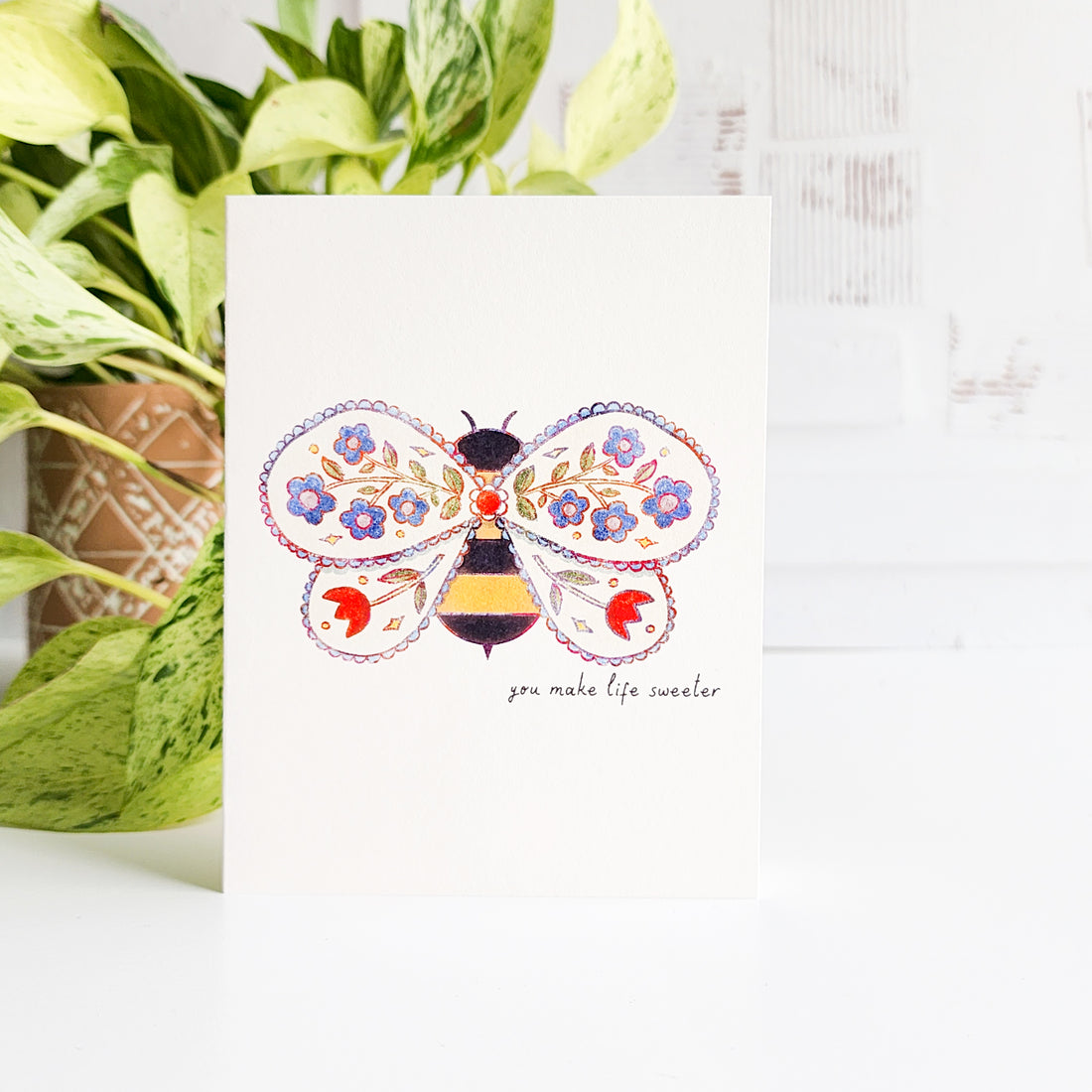 Greeting card with a decorative bee design and text, placed on a white surface with green plants.