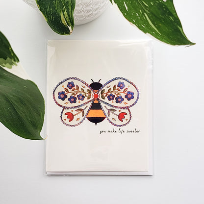 Card with a bee design with flowers on a white surface with a plant in the background