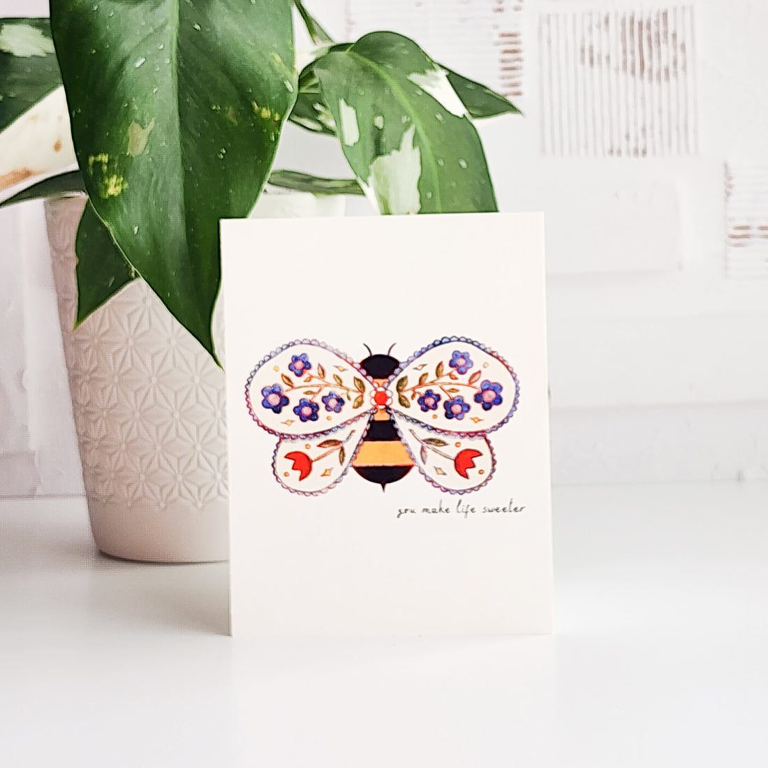Card with a bee design with flowers on a white surface with a plant in the background