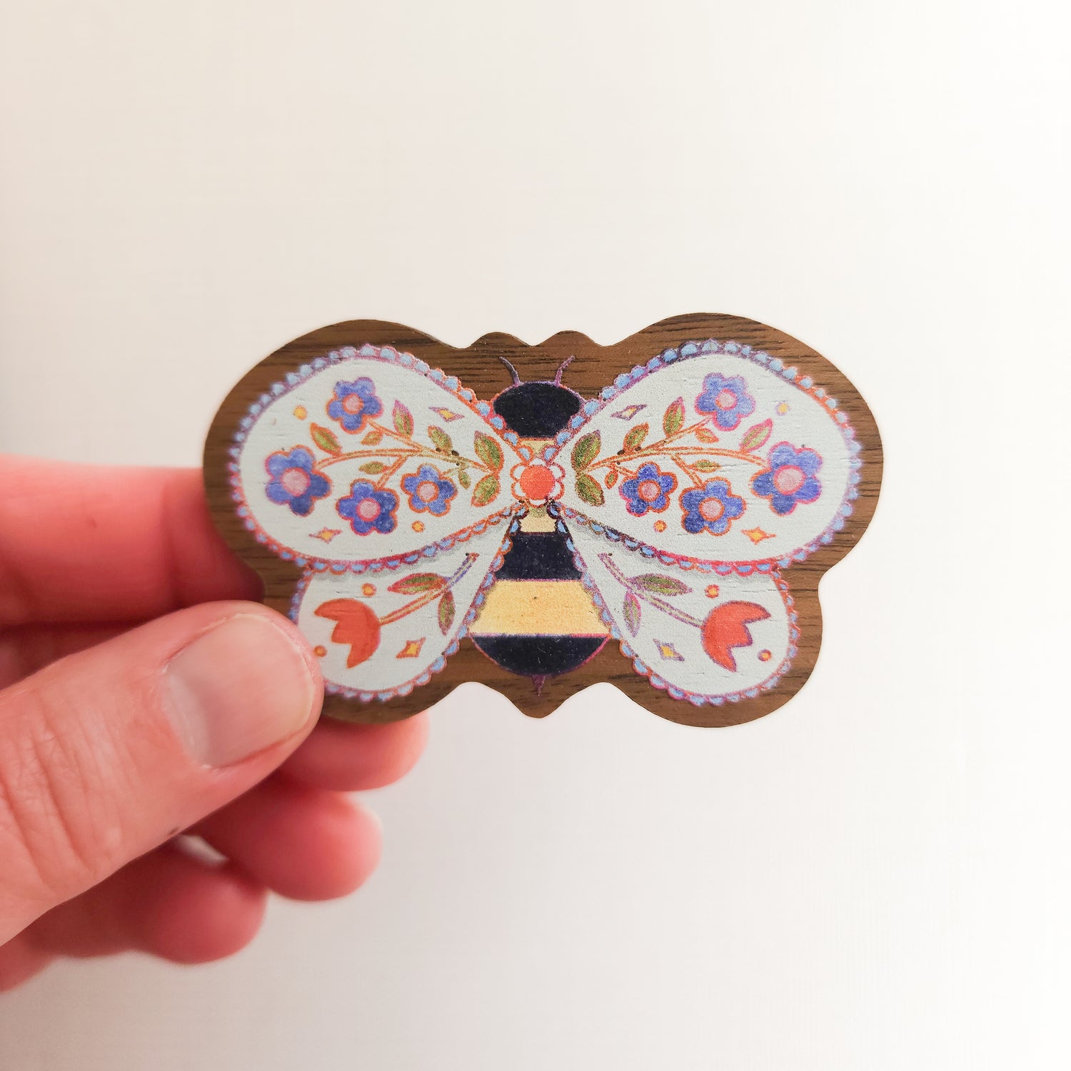 hand holding a wooden magnet with an illustration of a bee with flowers in its wings on a linen background