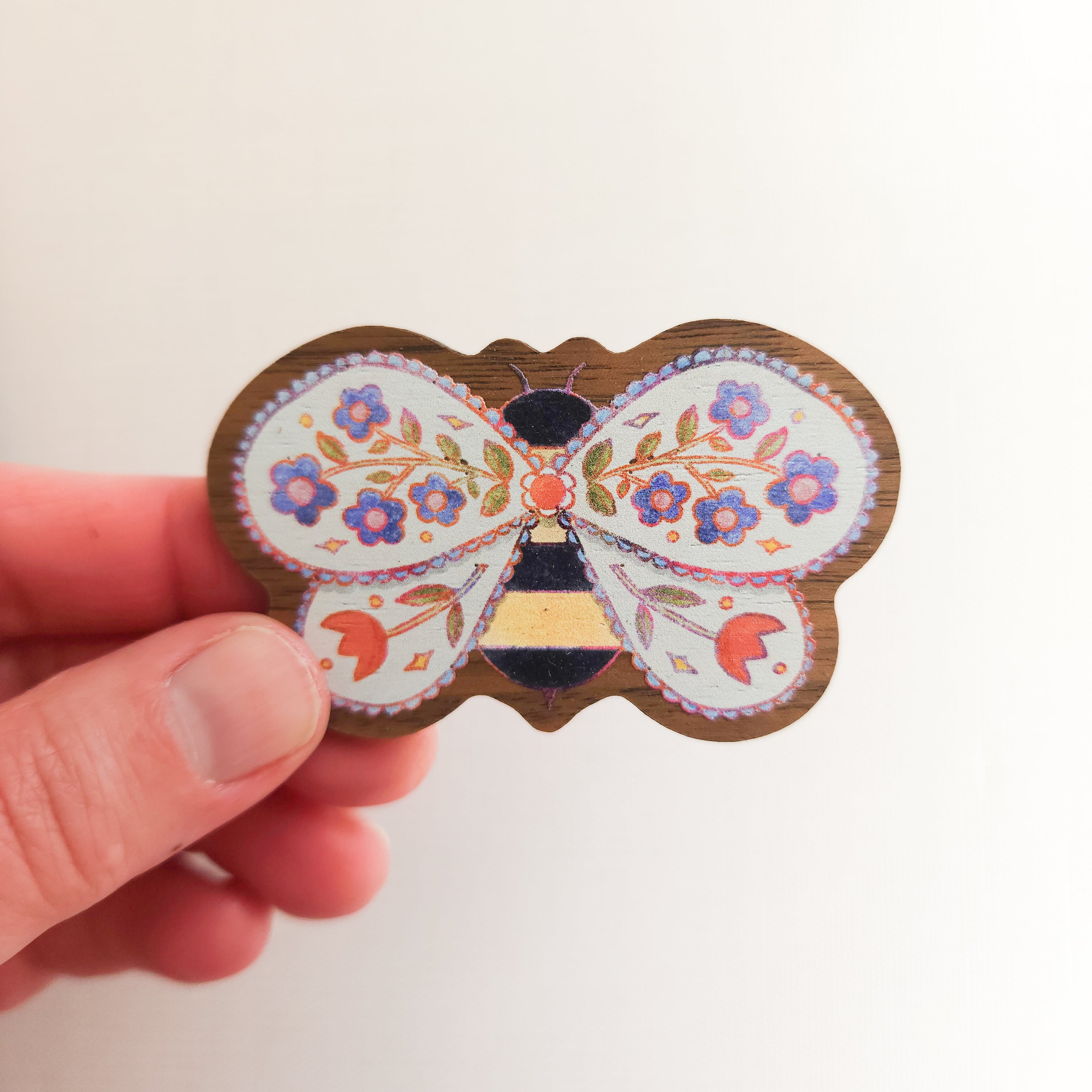 hand holding a wooden magnet with an illustration of a bee with flowers in its wings on a linen background
