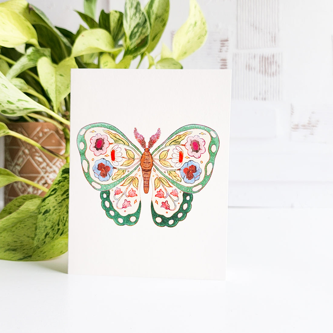 Butterfly greeting card card with floral design on a white surface with green plants.
