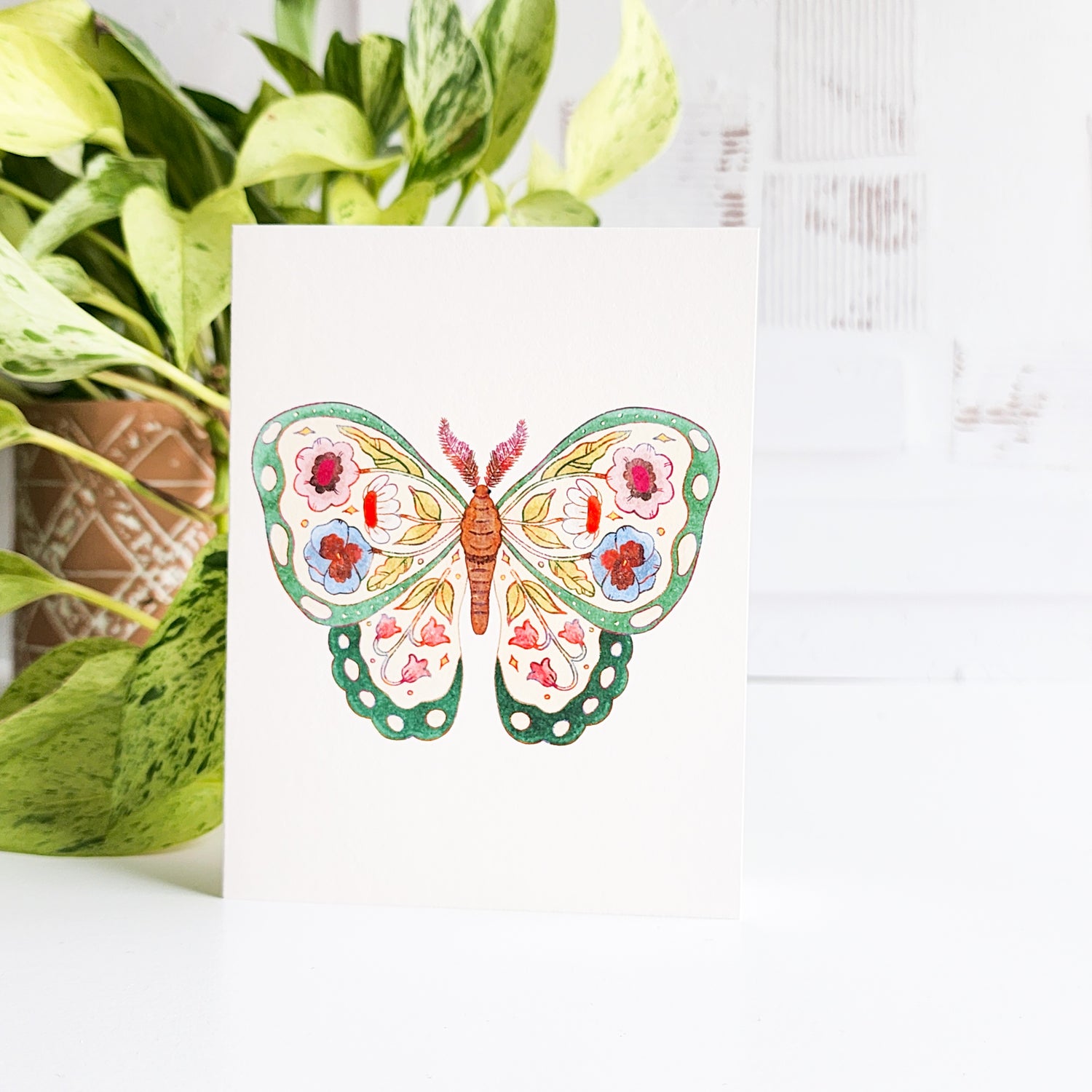 Butterfly greeting card card with floral design on a white surface with green plants.