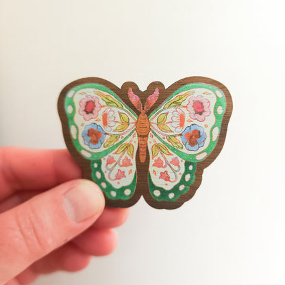 hand holding a wooden magnet with an illustration of a green butterfly with flowers in its wings