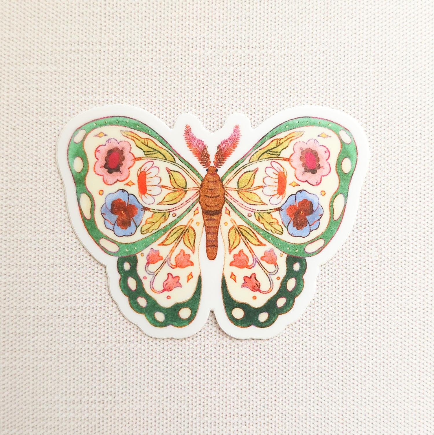 sticker of an illustrated green butterfly with flowers in its wings on a linen background