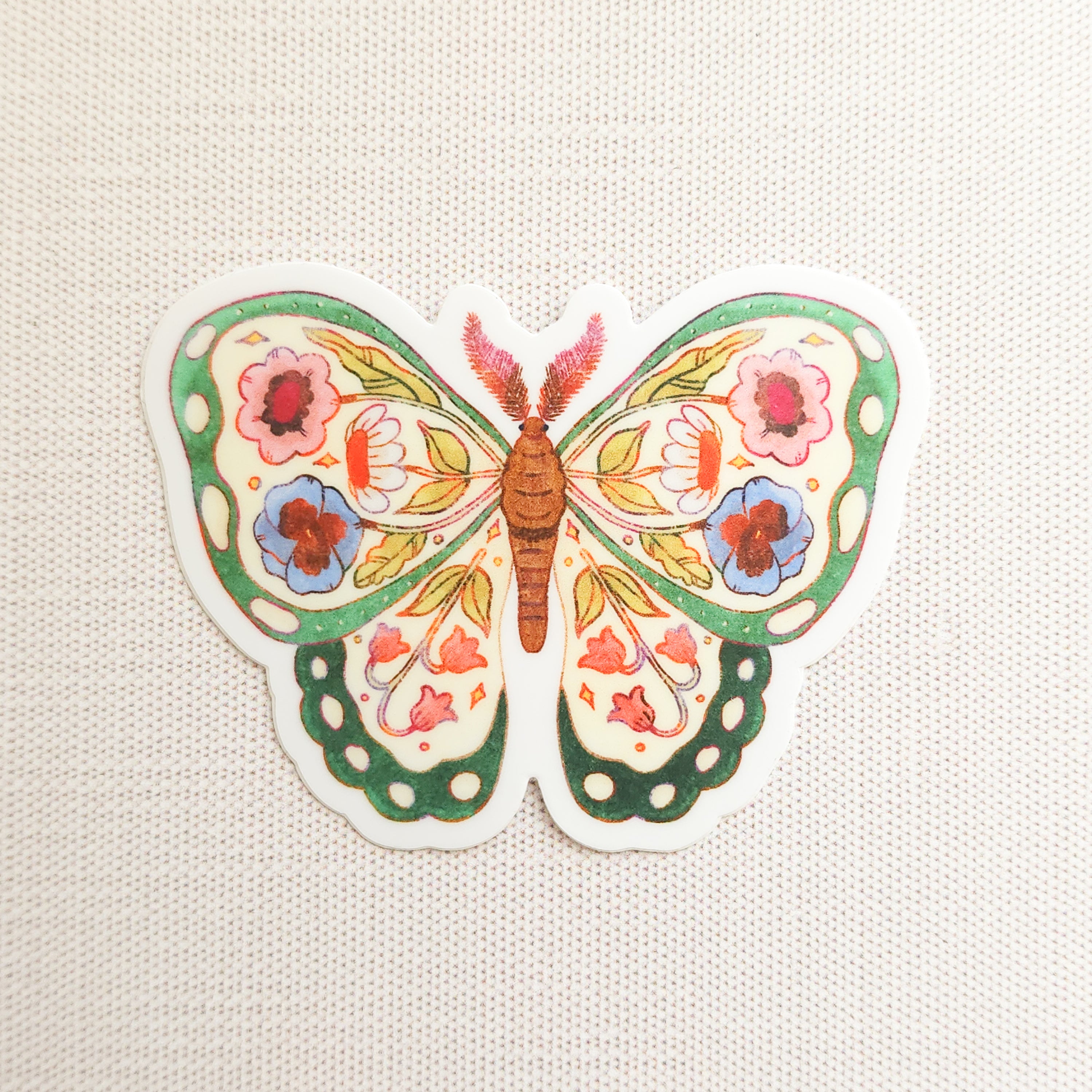 sticker of an illustrated green butterfly with flowers in its wings on a linen background