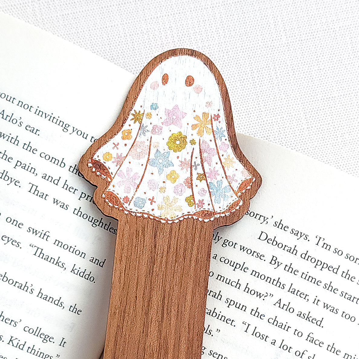 Wooden bookmark featuring a little floral ghost with colorful flowers and stars on its sheet, printed on walnut wood and photographed on an open book on a neutral fabric background.