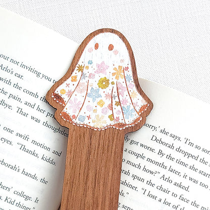 Wooden bookmark featuring a little floral ghost with colorful flowers and stars on its sheet, printed on walnut wood and photographed on an open book on a neutral fabric background.