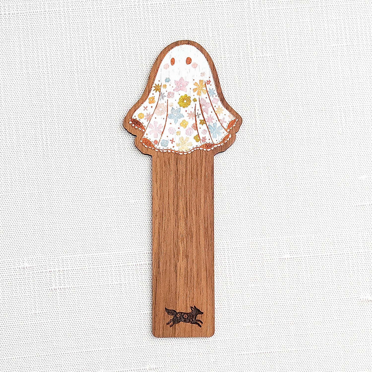 Wooden bookmark featuring a little floral ghost with colorful flowers and stars on its sheet, printed on walnut wood and photographed on a neutral fabric background.