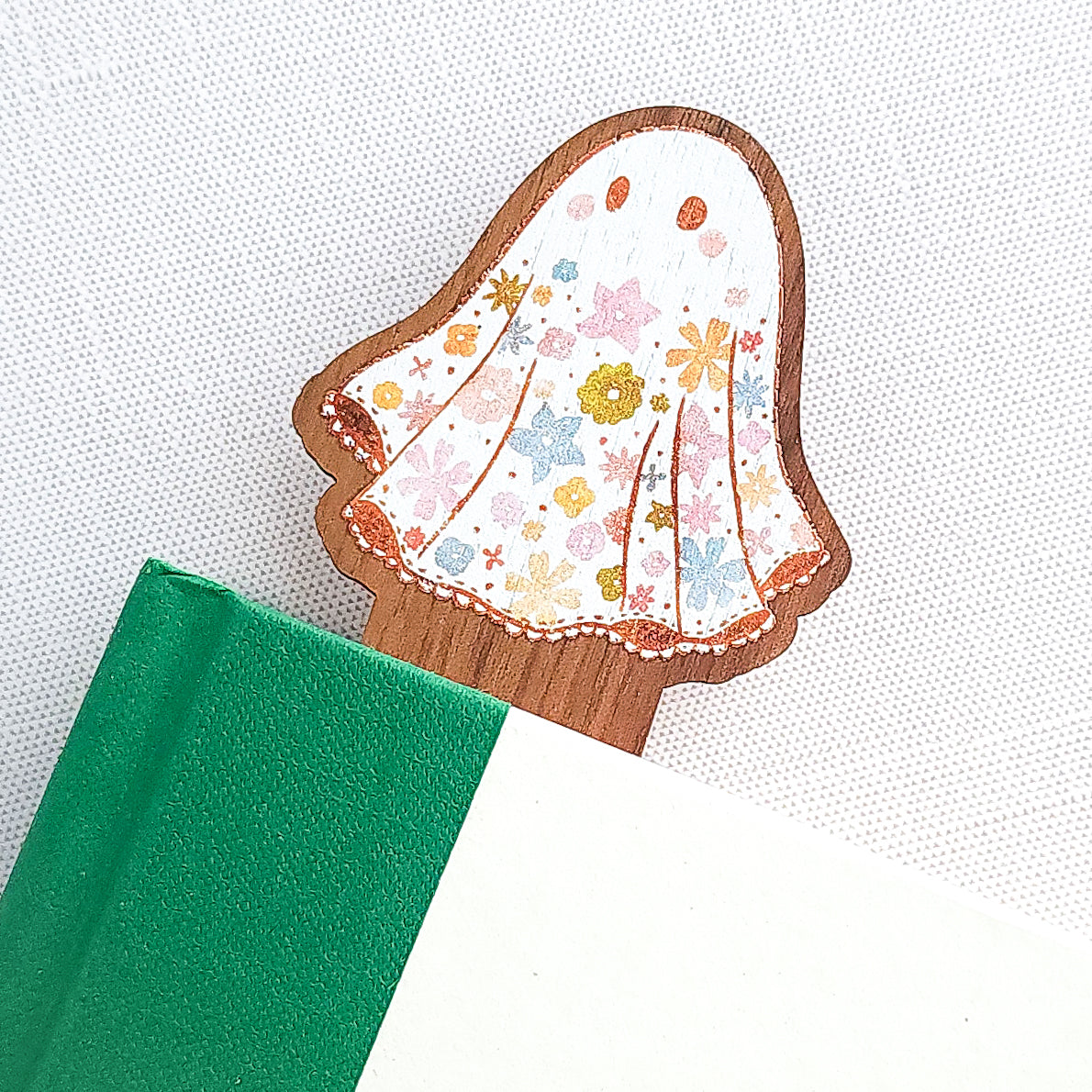 Wooden bookmark featuring a little floral ghost with colorful flowers and stars on its sheet, printed on walnut wood and photographed inside a book on a neutral fabric background.