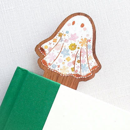 Wooden bookmark featuring a little floral ghost with colorful flowers and stars on its sheet, printed on walnut wood and photographed inside a book on a neutral fabric background.