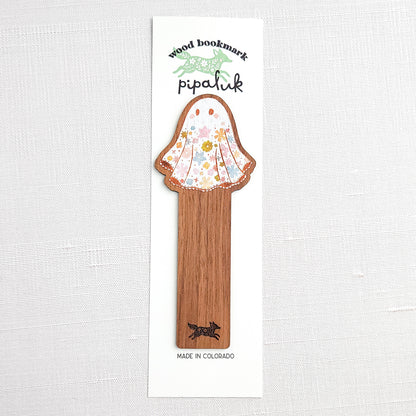 Wooden bookmark featuring a little floral ghost with colorful flowers and stars on its sheet, printed on walnut wood and photographed in Pipaluk branded packaging on a neutral fabric background