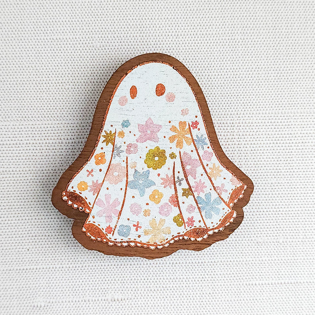Wooden magnet featuring a little floral ghost with colorful flowers and stars on its sheet, printed on walnut wood and photographed on a neutral fabric background.
